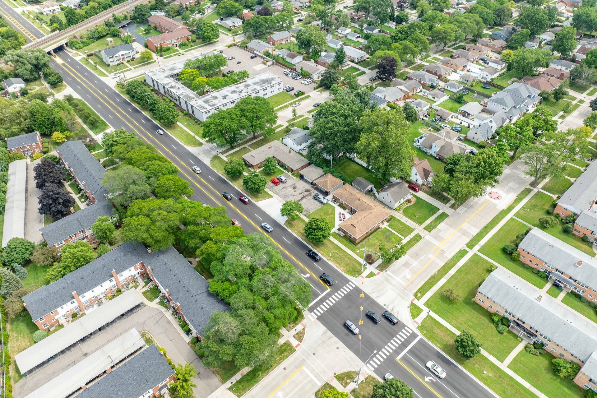 Aerial view of a suburban neighborhood with a wide road, trees, houses, and cars driving on the street.