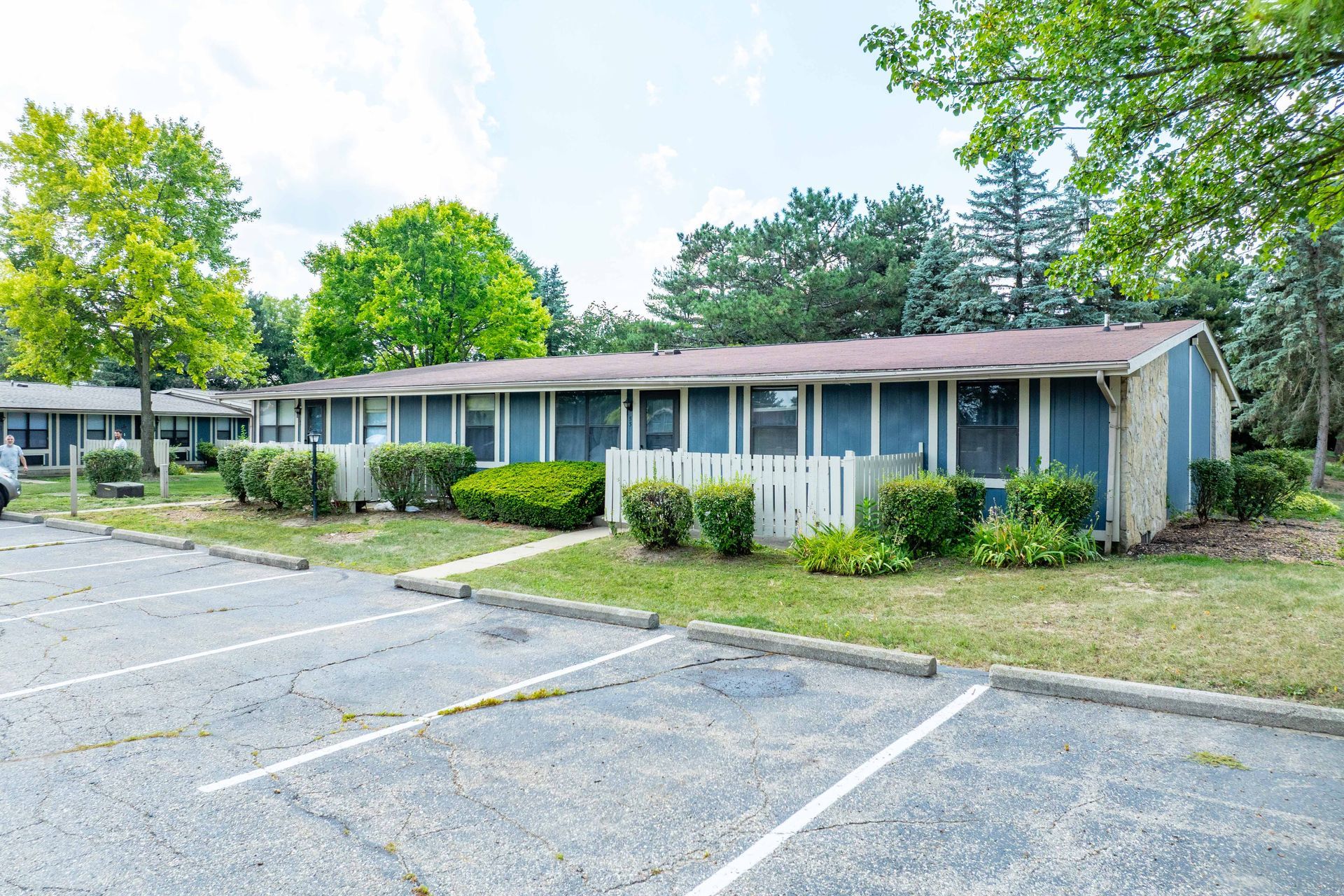 Blue apartment building with parking lot, white fence, green bushes, and trees on a sunny day.