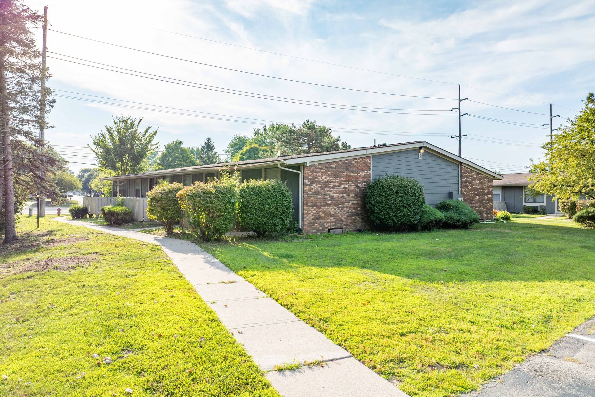 Sidewalk leading to a one-story brick building with green bushes and grass on a sunny day.