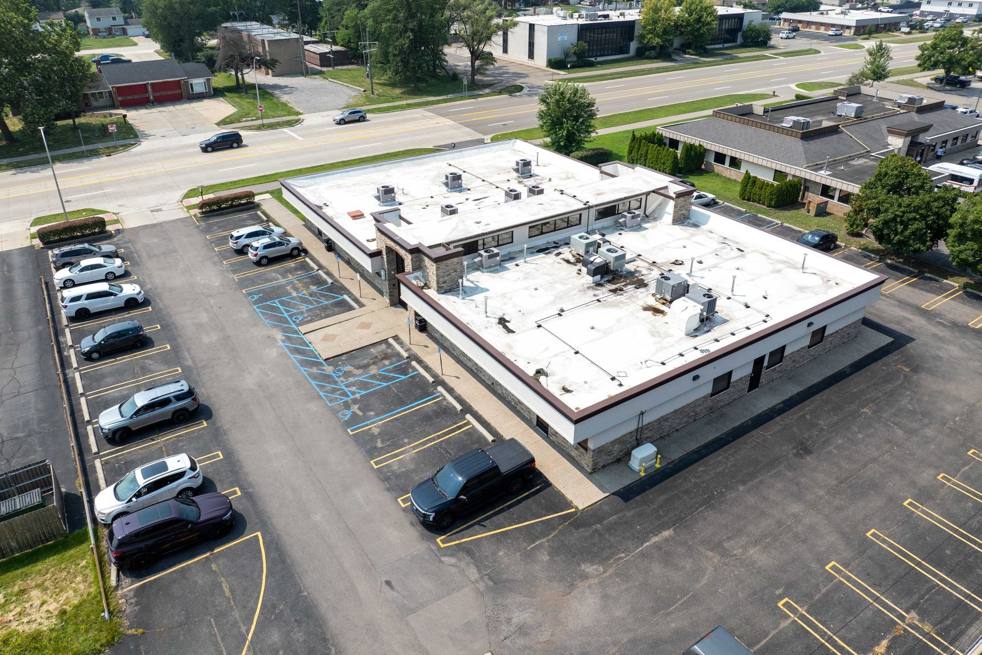 Aerial view of a commercial building with a white roof, surrounded by parking spaces. Cars are parked in designated spots.