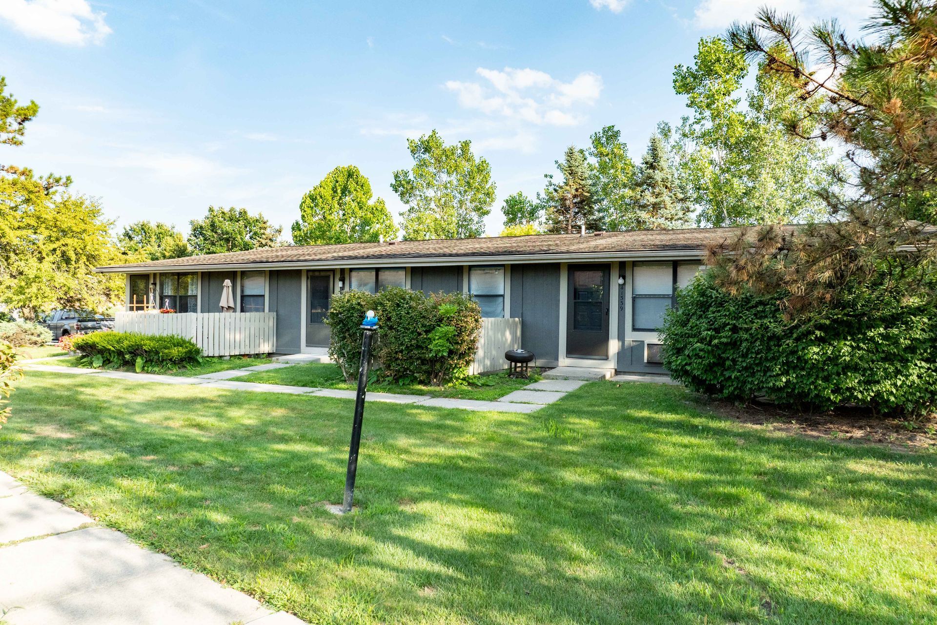 Two gray, single-story apartments with grassy lawn under a blue sky and trees.