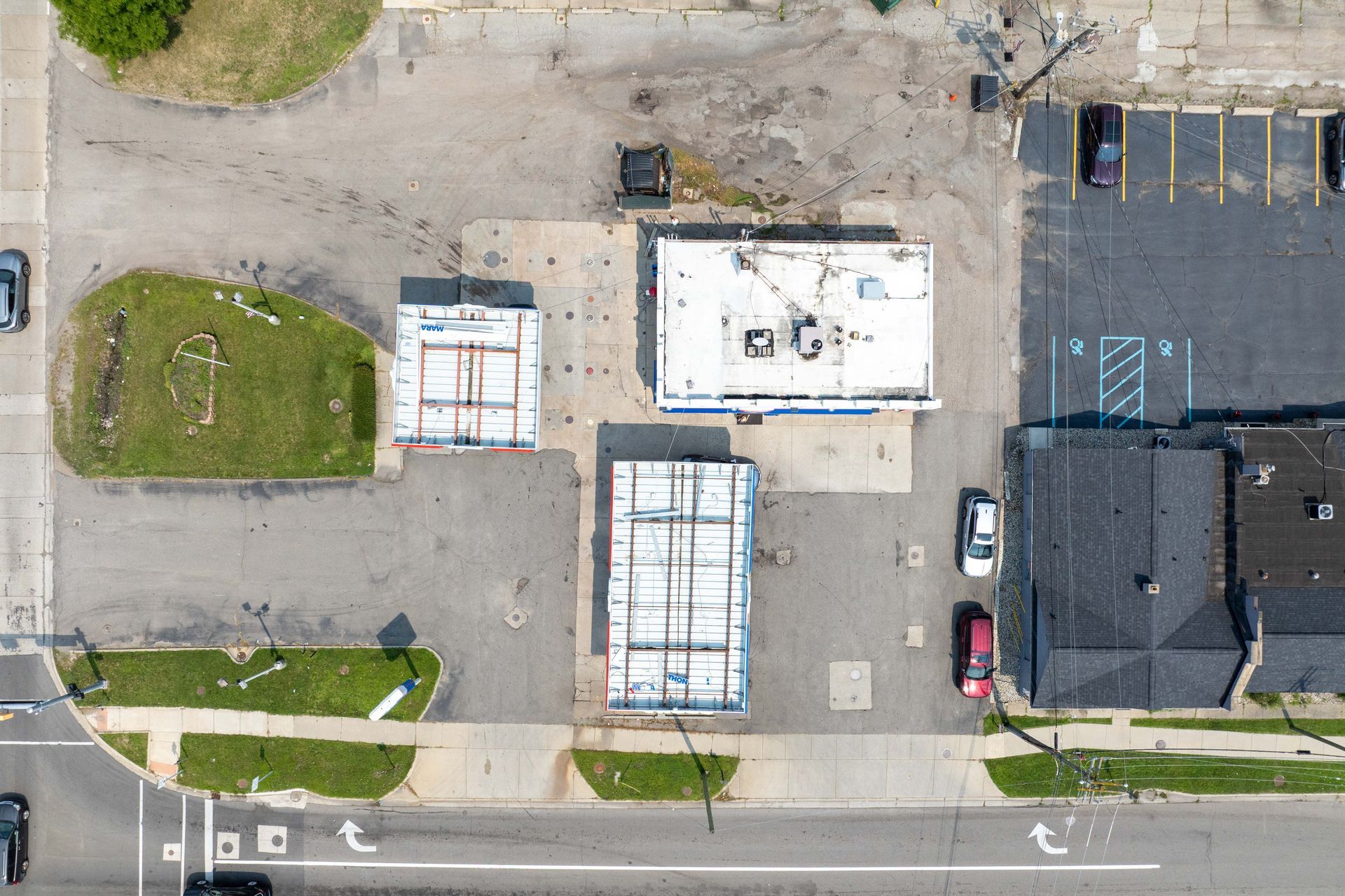 Overhead view of buildings with parking areas. Two metal sheds near building with white roof. Road and greenery below.