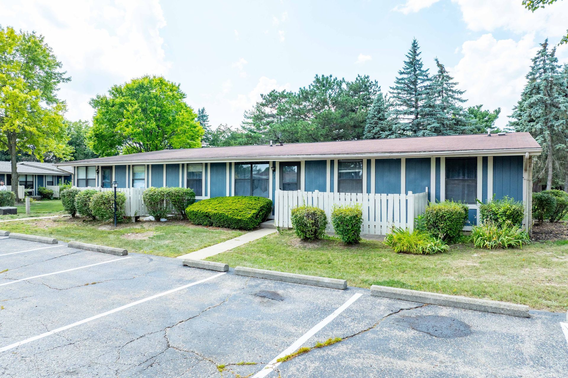 Low-rise, blue apartment building with white fence, green grass, and parking spaces.