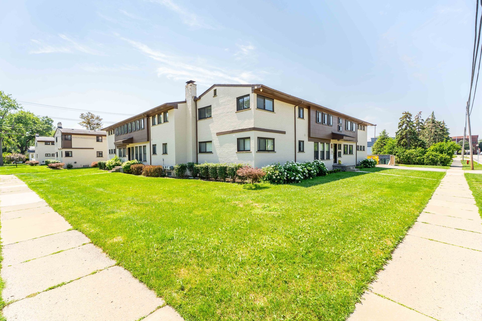 Multi-unit building with tan exterior, green lawn, sidewalk, and trees.