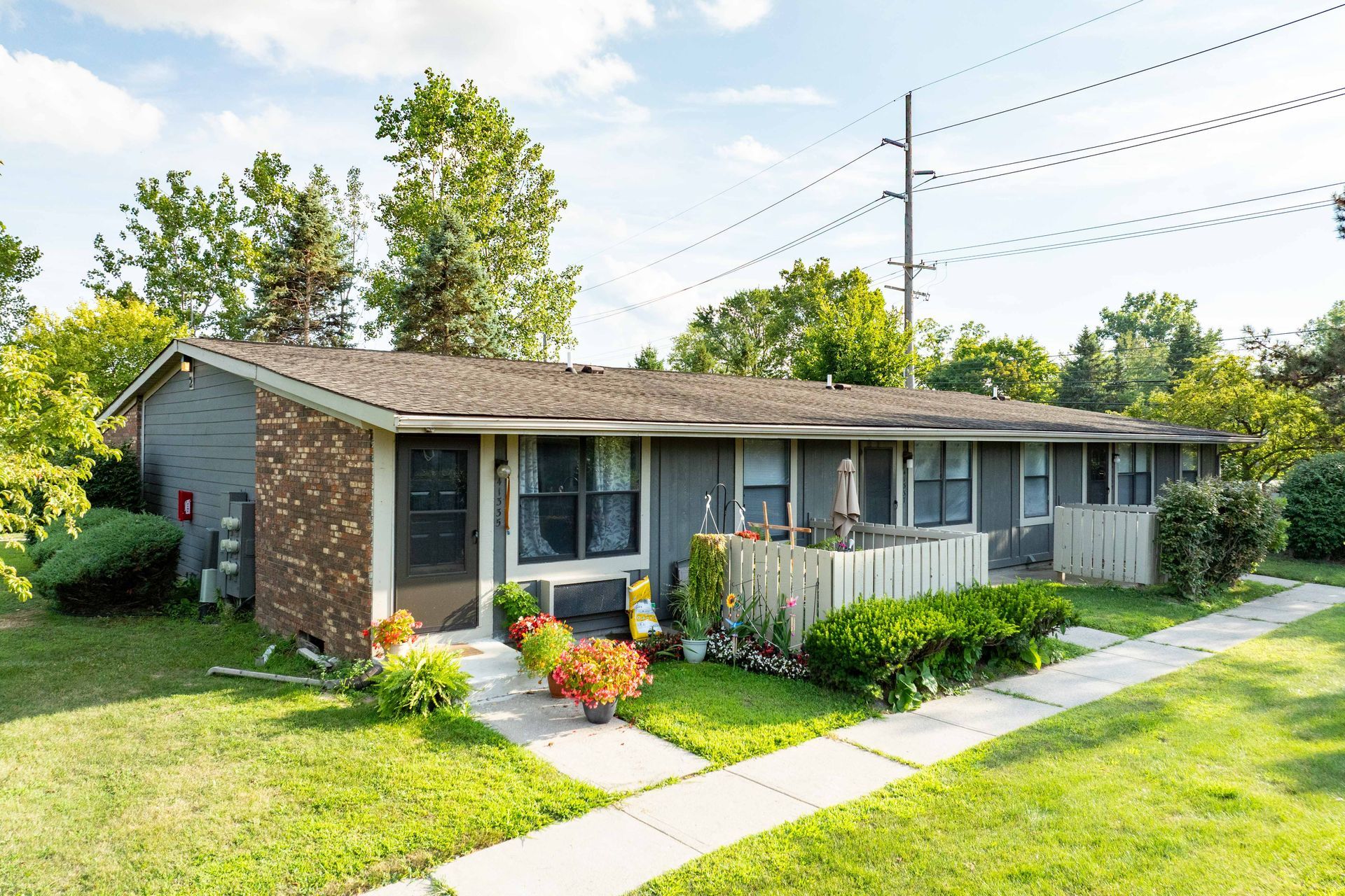 A one-story gray building with a brick facade, featuring a walkway, greenery, and overhead power lines.