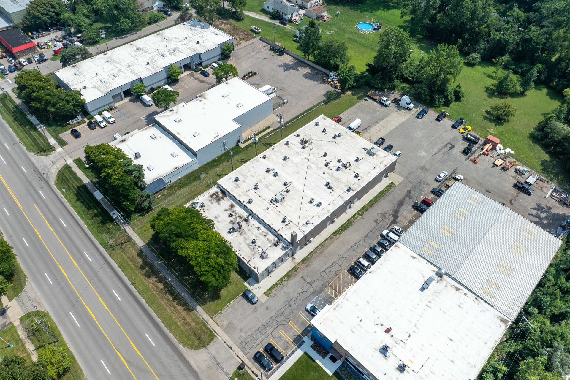 Aerial view of several industrial buildings with white roofs and parking lots, surrounded by trees and a road.