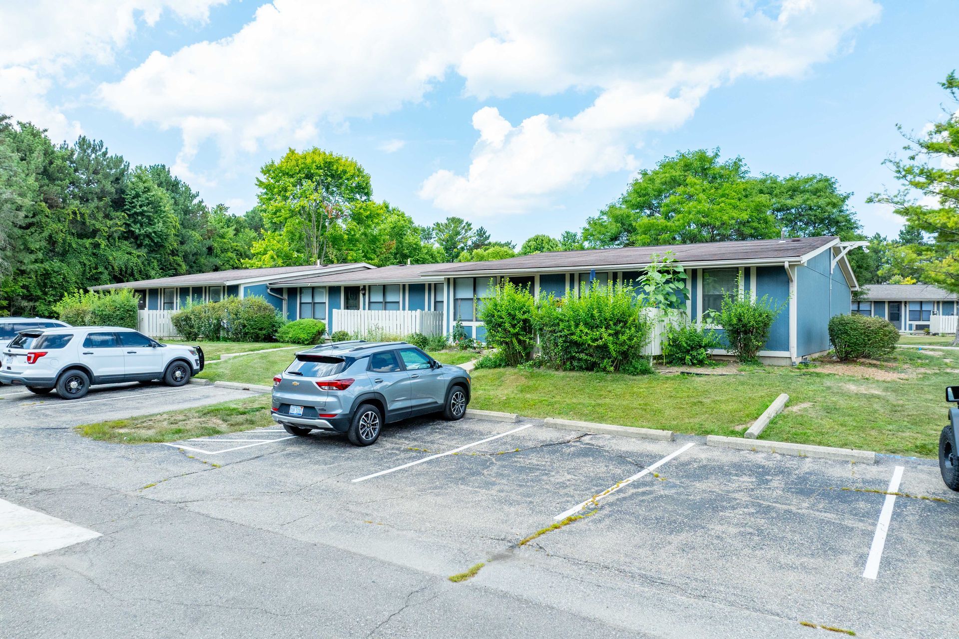 Exterior view of blue apartment buildings with parked cars in a parking lot on a sunny day.