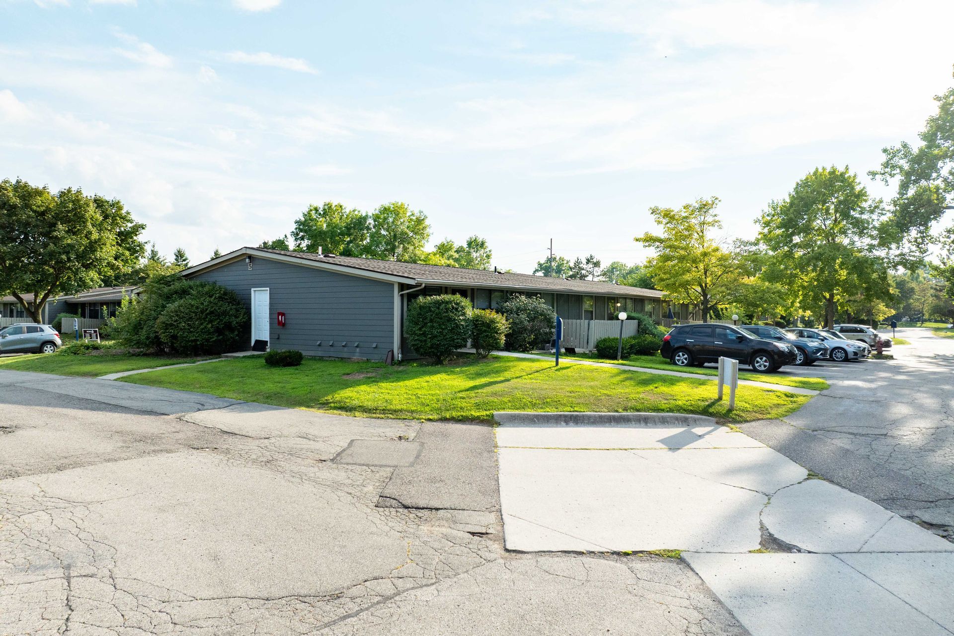 Gray building with parked cars, grassy area, and paved road on a sunny day.