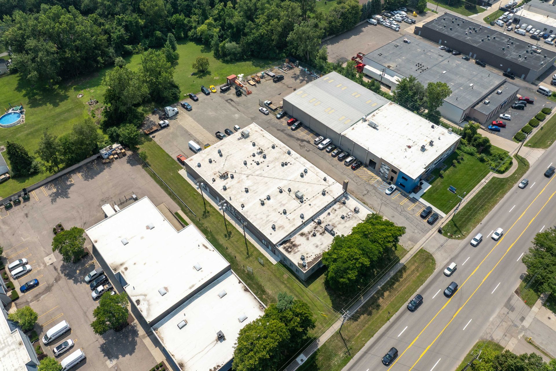Aerial view of industrial buildings with parking lots, trees, and a road with cars.