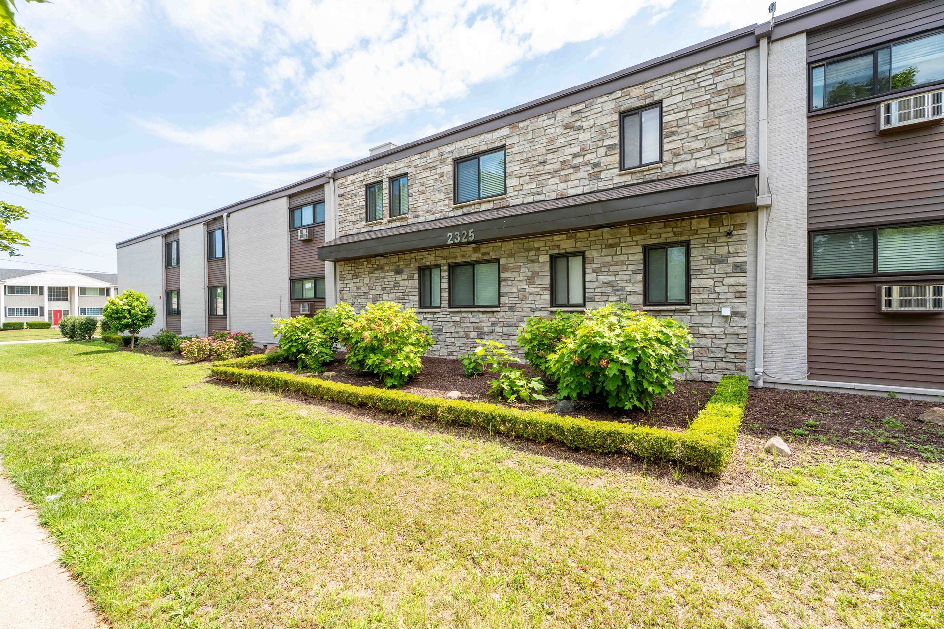 Apartment building exterior with stone facade, green lawn, and bushes in front.