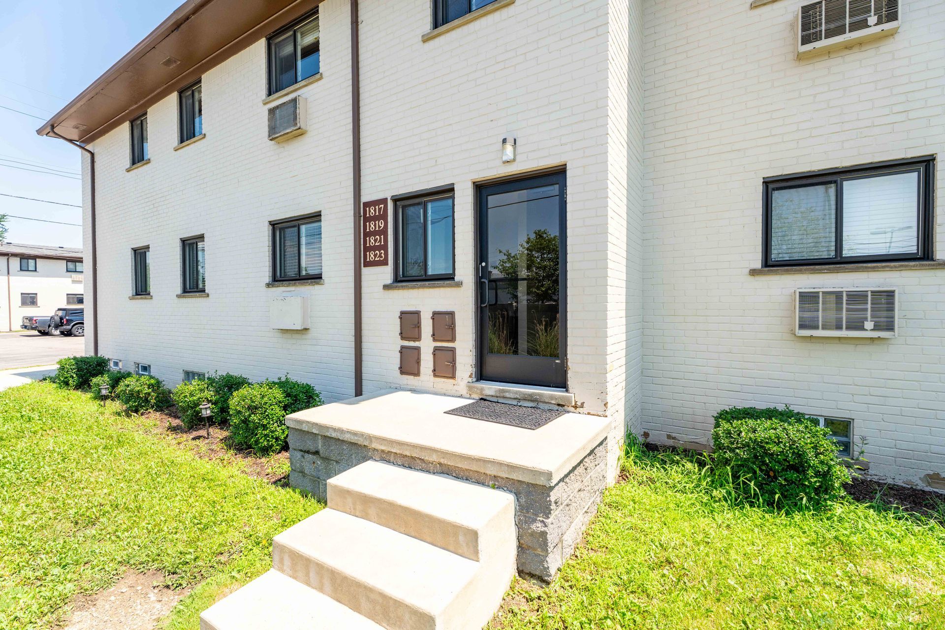 Two-story brick apartment building with black door and steps. Green bushes line the lawn in front.