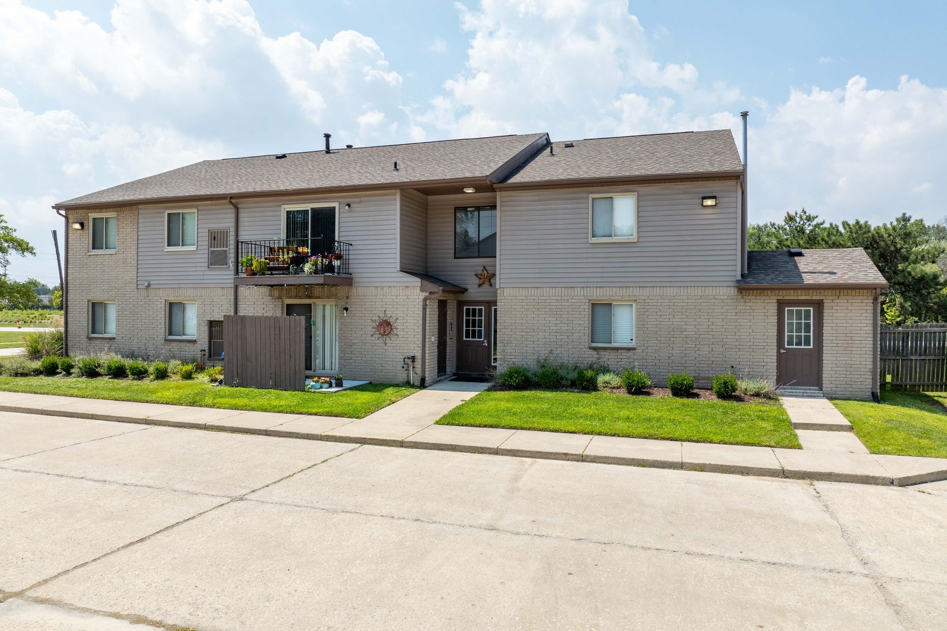 Two-story apartment building with tan brick and siding, a balcony with plants, and a grassy lawn.