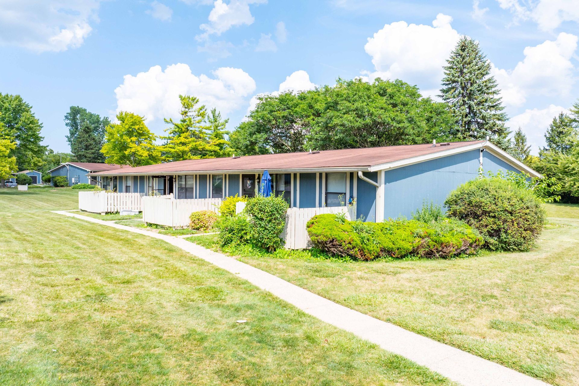 Blue-sided, one-story apartment buildings with white accents, a walkway, and green lawn under a partly cloudy sky.