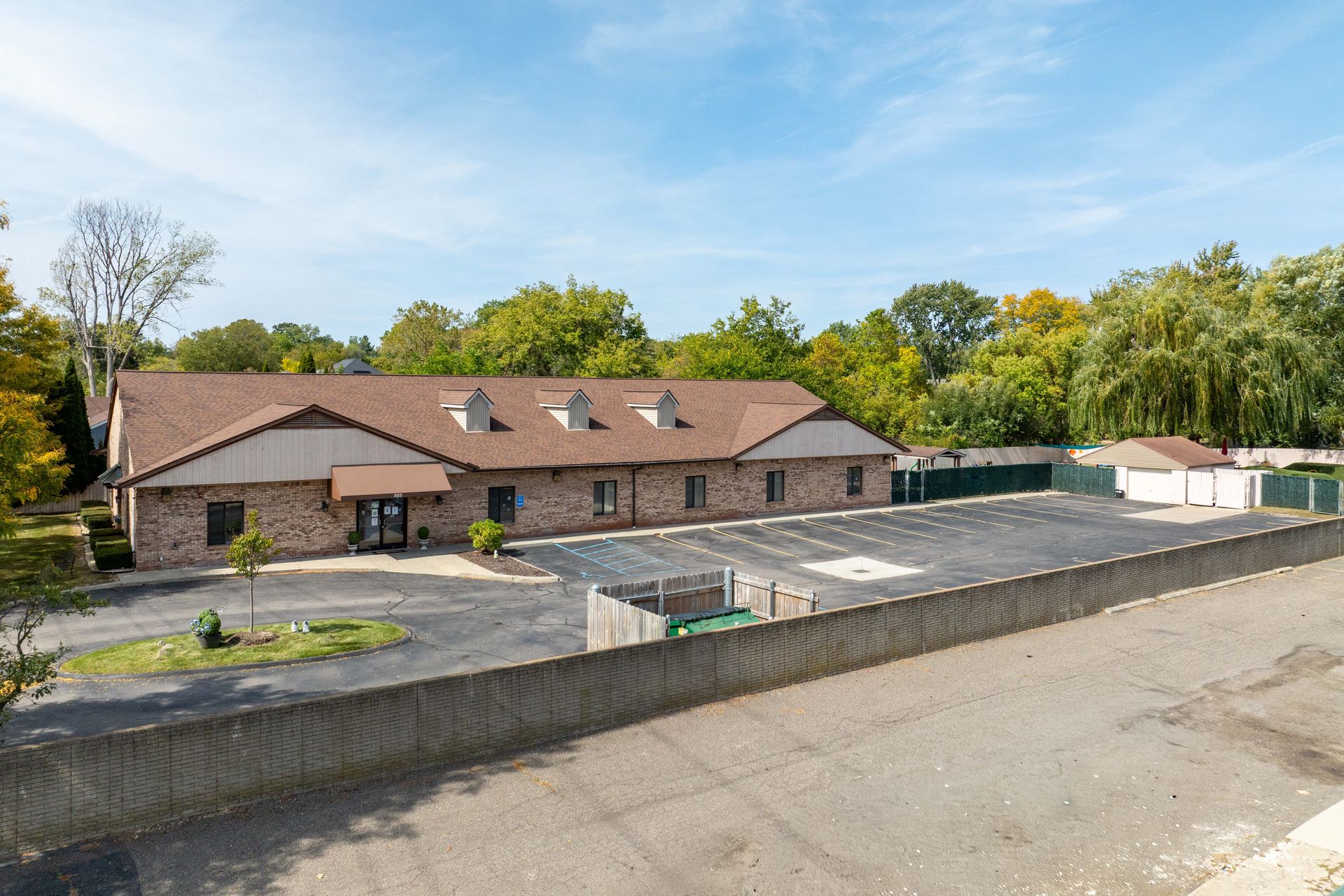 Low-angle view of a brick building with a brown roof and a black parking lot, with surrounding trees and a chain-link fence.