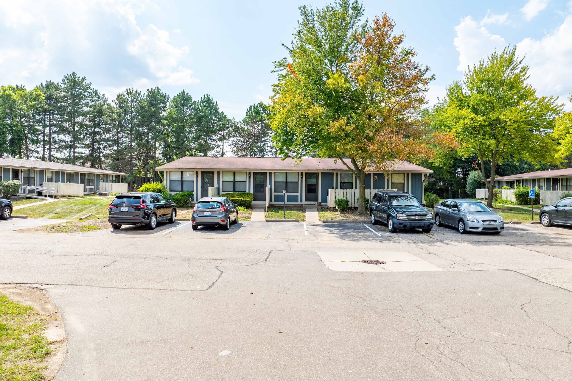 Apartment complex with cars parked in front. Gray buildings with green trees and a blue sky.