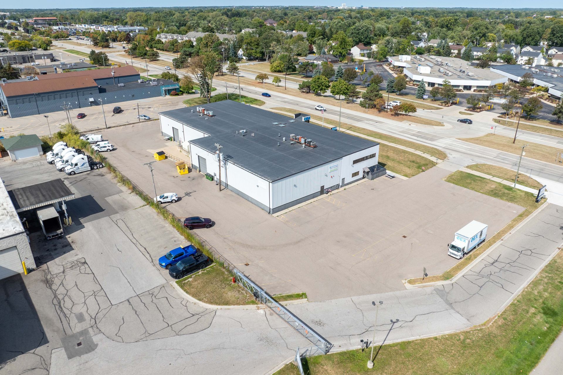 Aerial view of a white commercial building with a black roof, parking lot, and nearby roads and green areas.