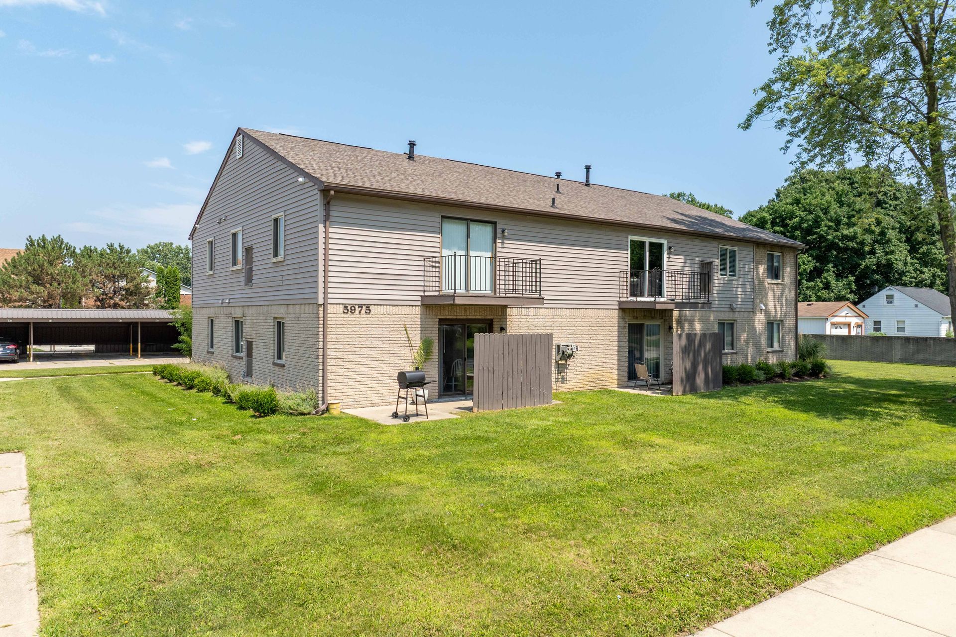 Two-story beige apartment building with green lawn and blue sky; a covered parking area on the left.
