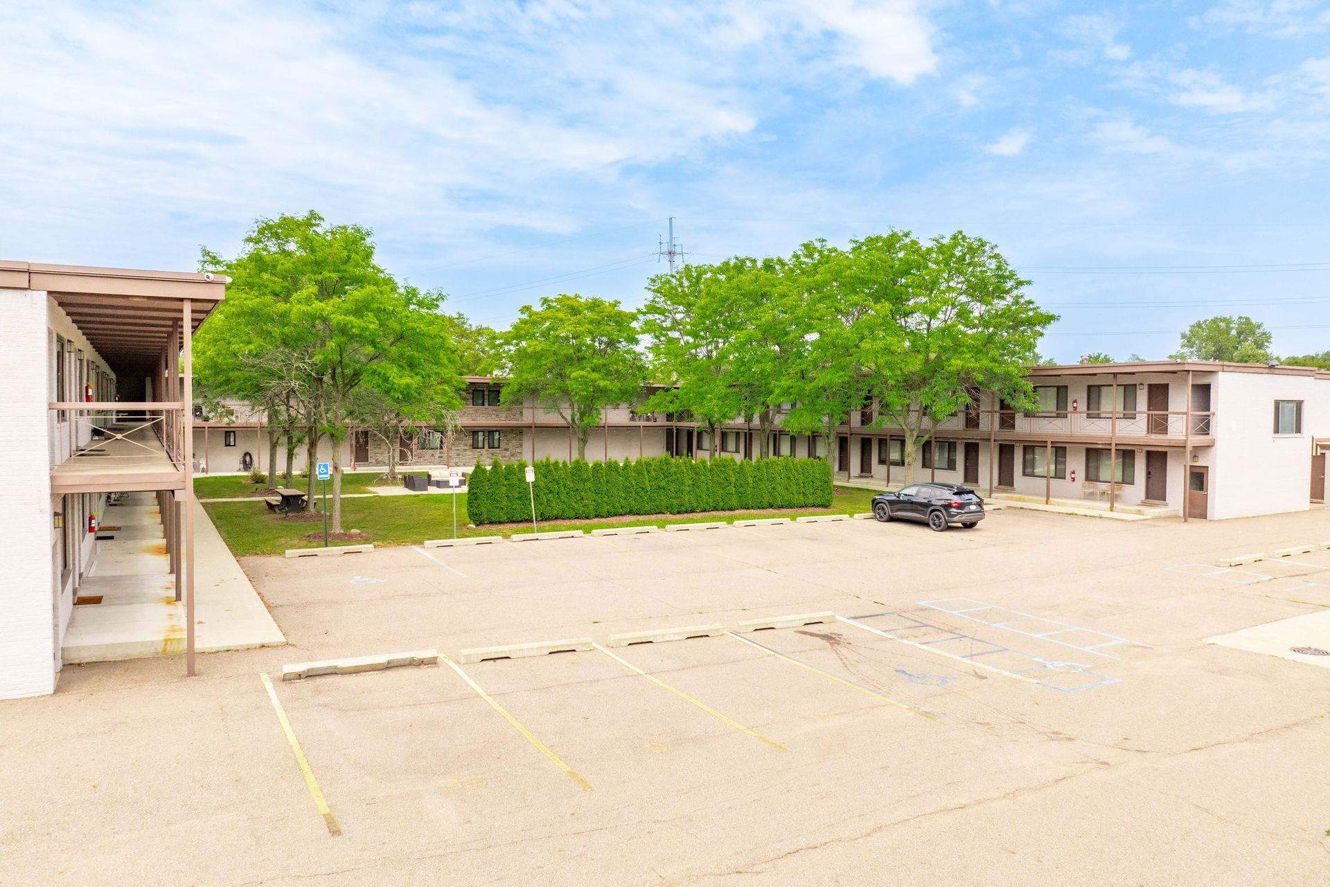 Exterior view of a motel with a parking lot in front. Buildings are tan with trees and blue sky.