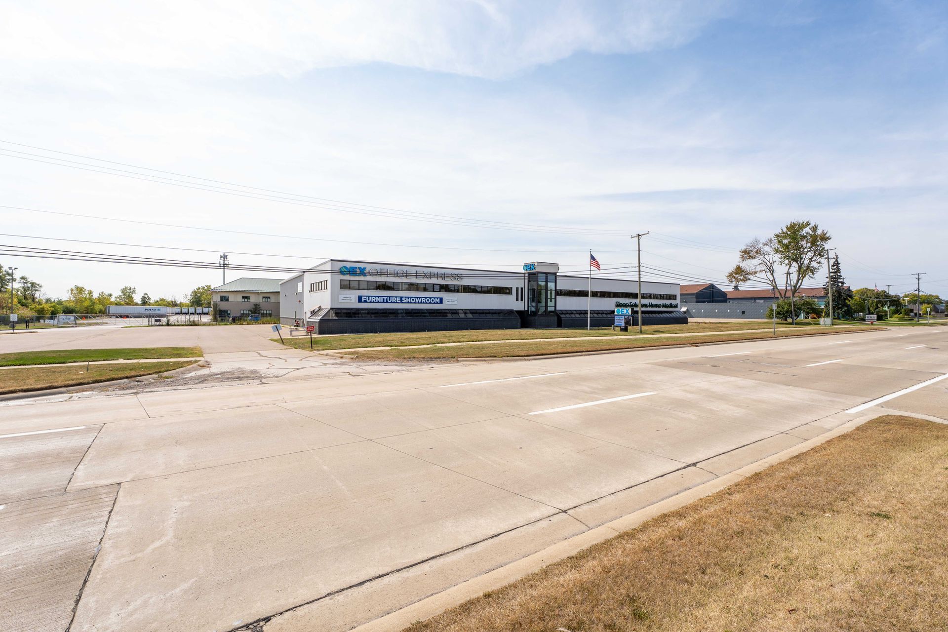 Two-story commercial building with blue accents, set on a grassy lot beside a paved road under a blue sky.