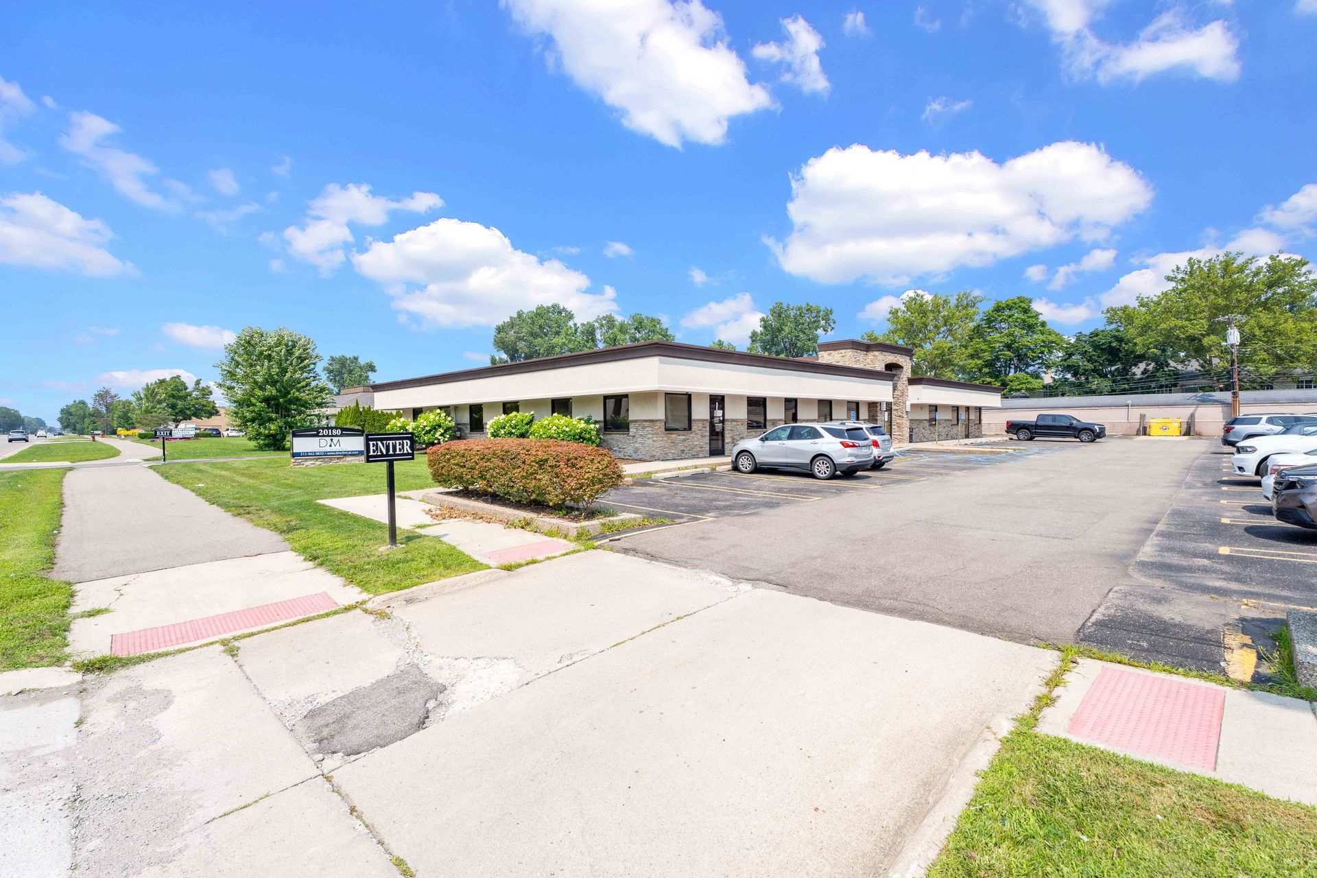 Low, tan office building with parking lot.  Cars, green bushes, sidewalk, and blue sky visible.