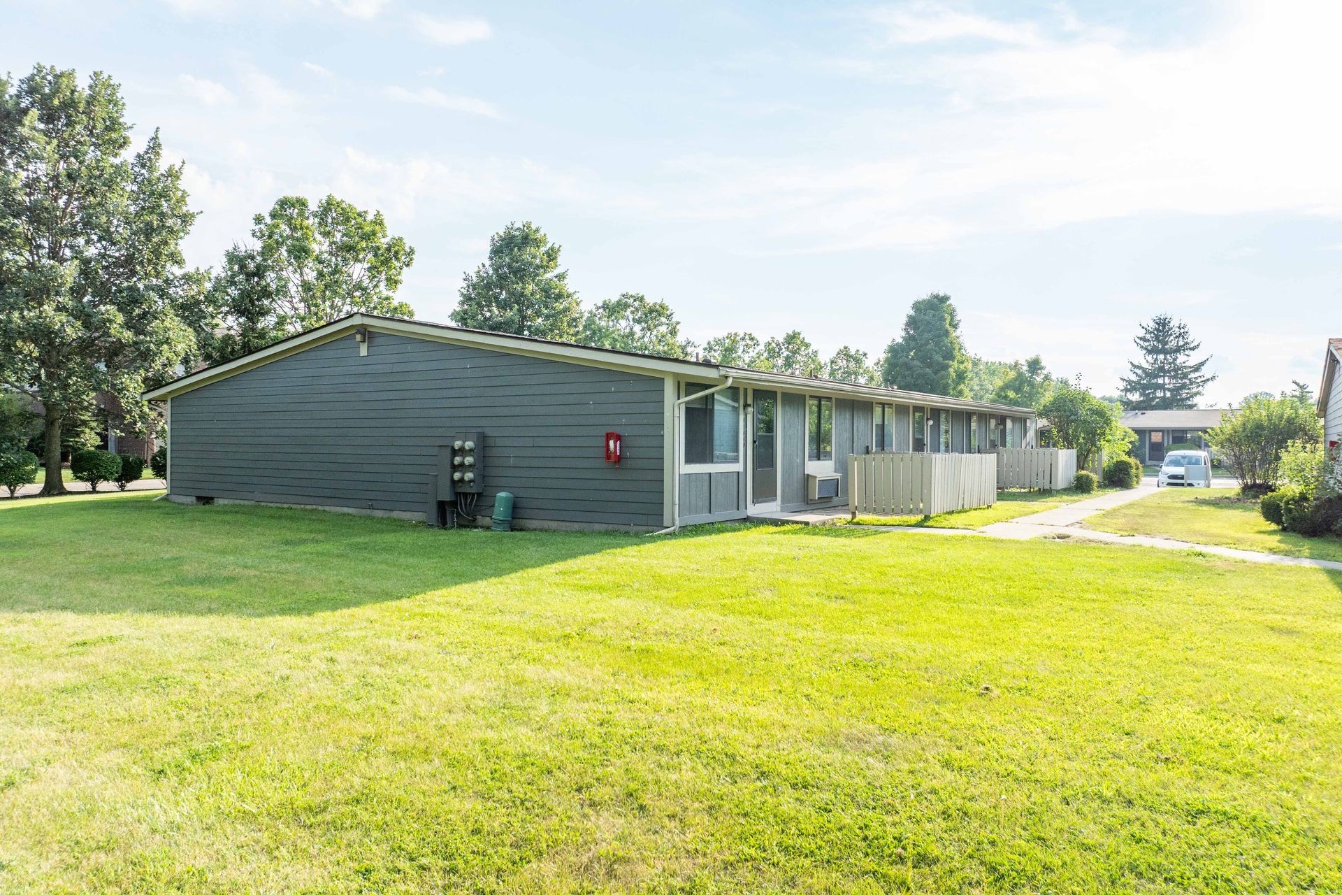 A one-story green building with a grassy yard and trees under a sunny sky.