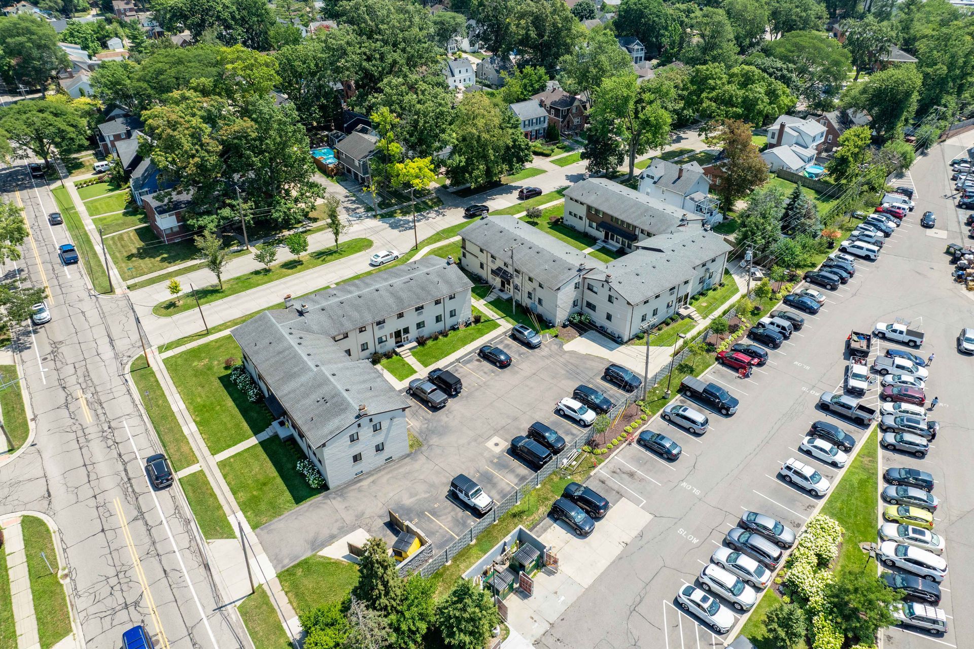 Aerial view of white apartment buildings with parking lot full of cars and surrounded by green trees.