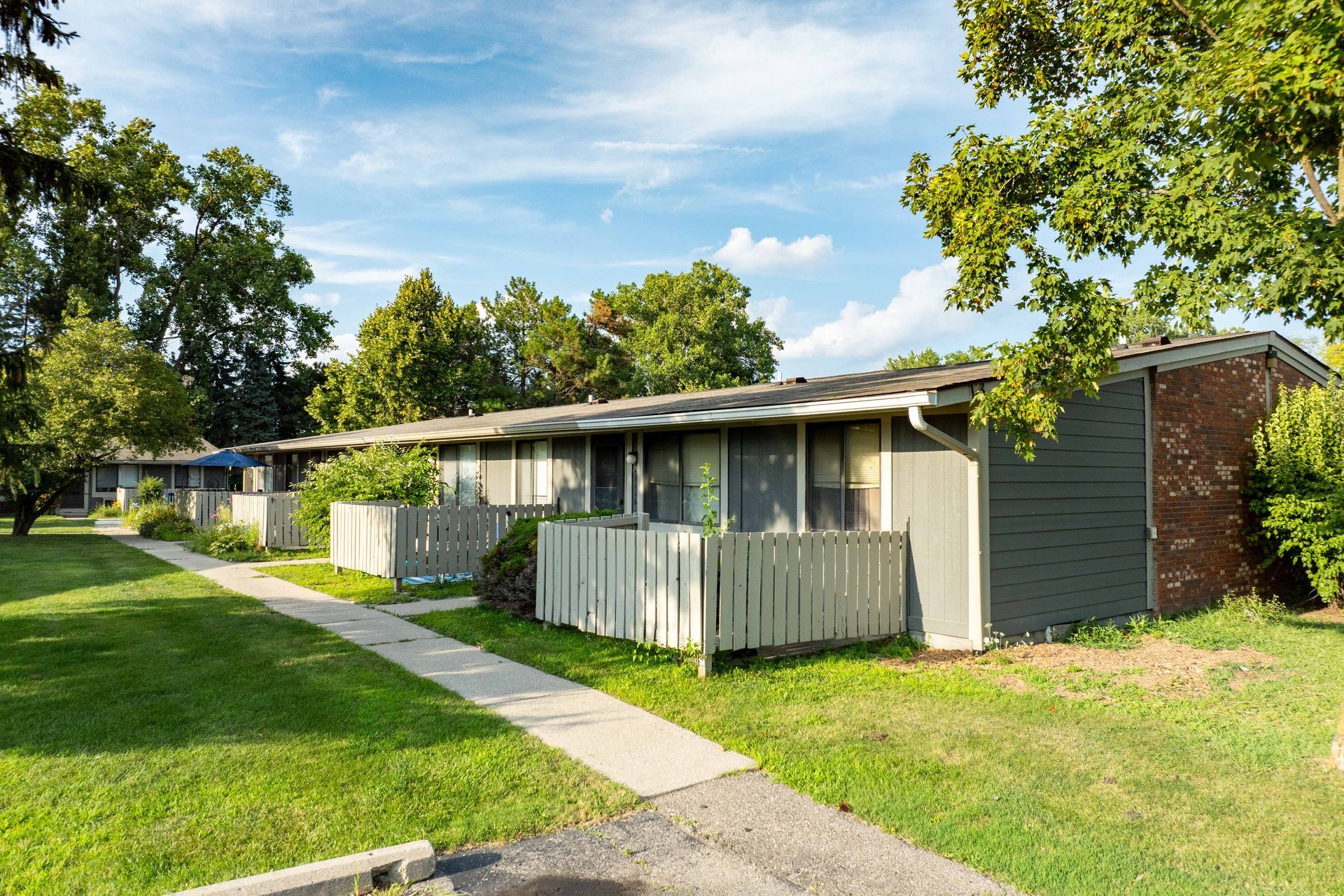 Row of apartments with green grass, walkway, and trees under a blue sky.