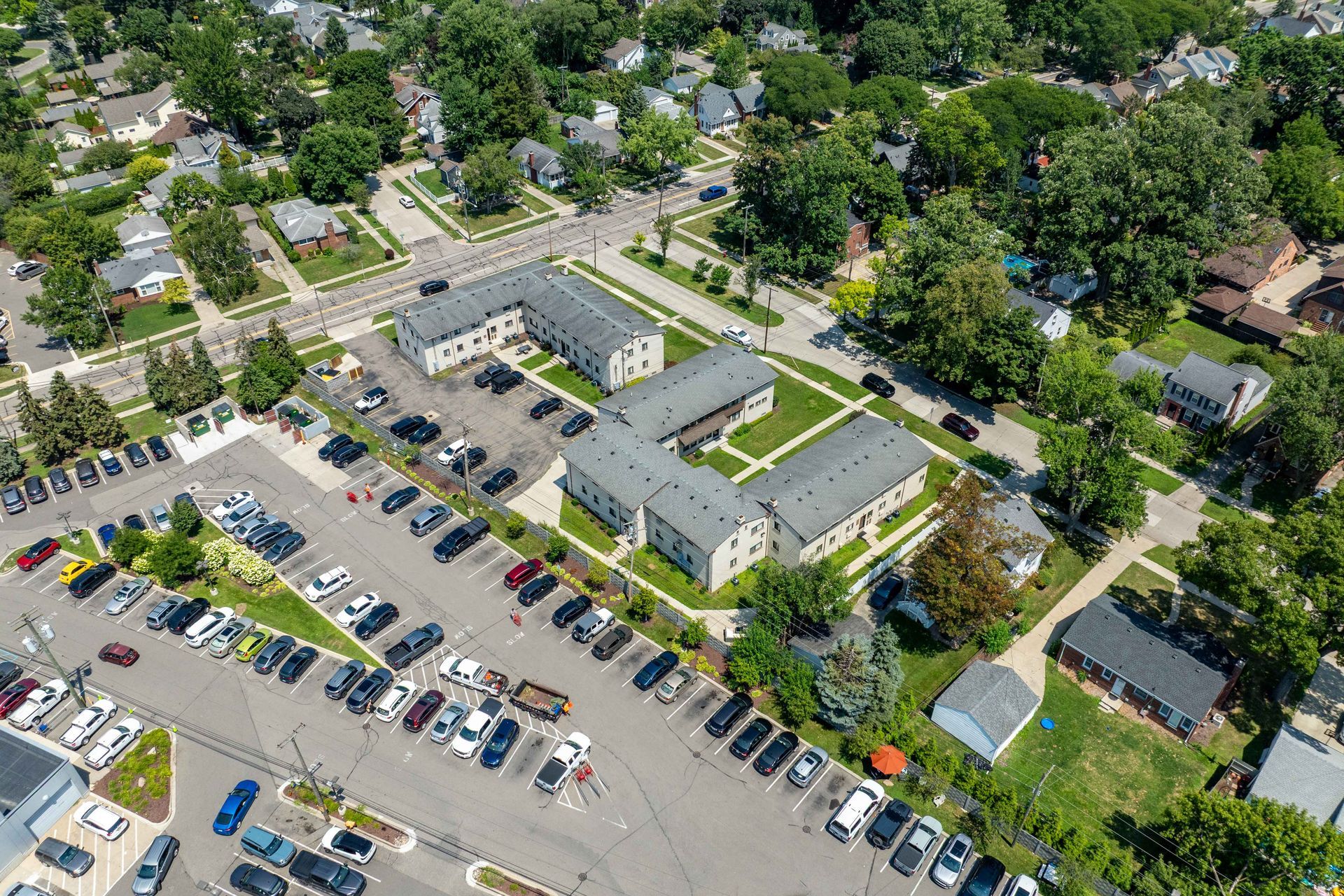Aerial view of apartment buildings with a large parking lot, surrounded by trees and houses in a residential area.