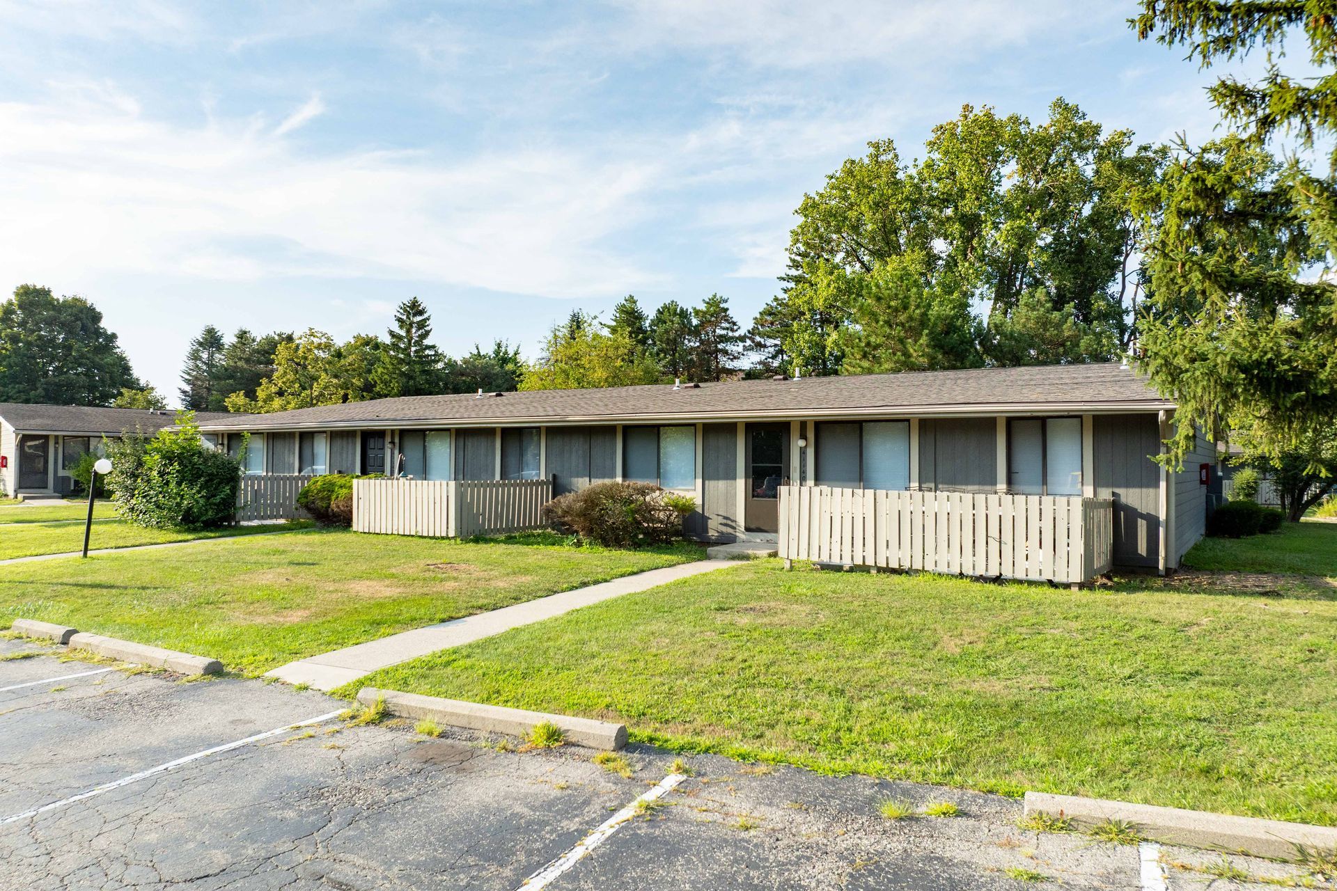 Low-rise apartment buildings with beige siding and fenced patios sit on green lawns under a blue sky.