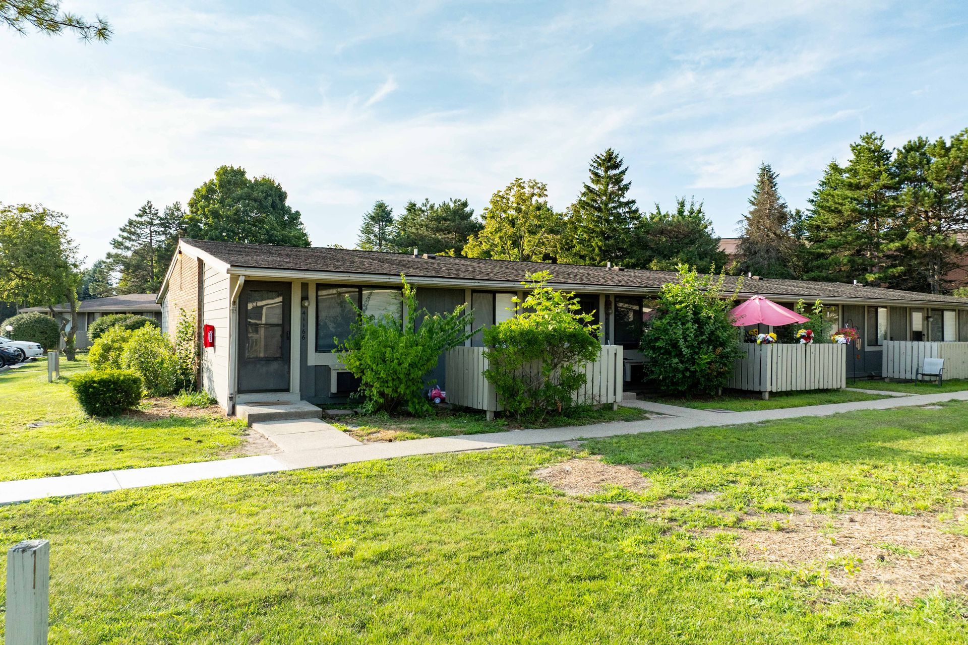 Low-rise apartments with grassy yard and trees under blue sky. A pink umbrella shades a patio.