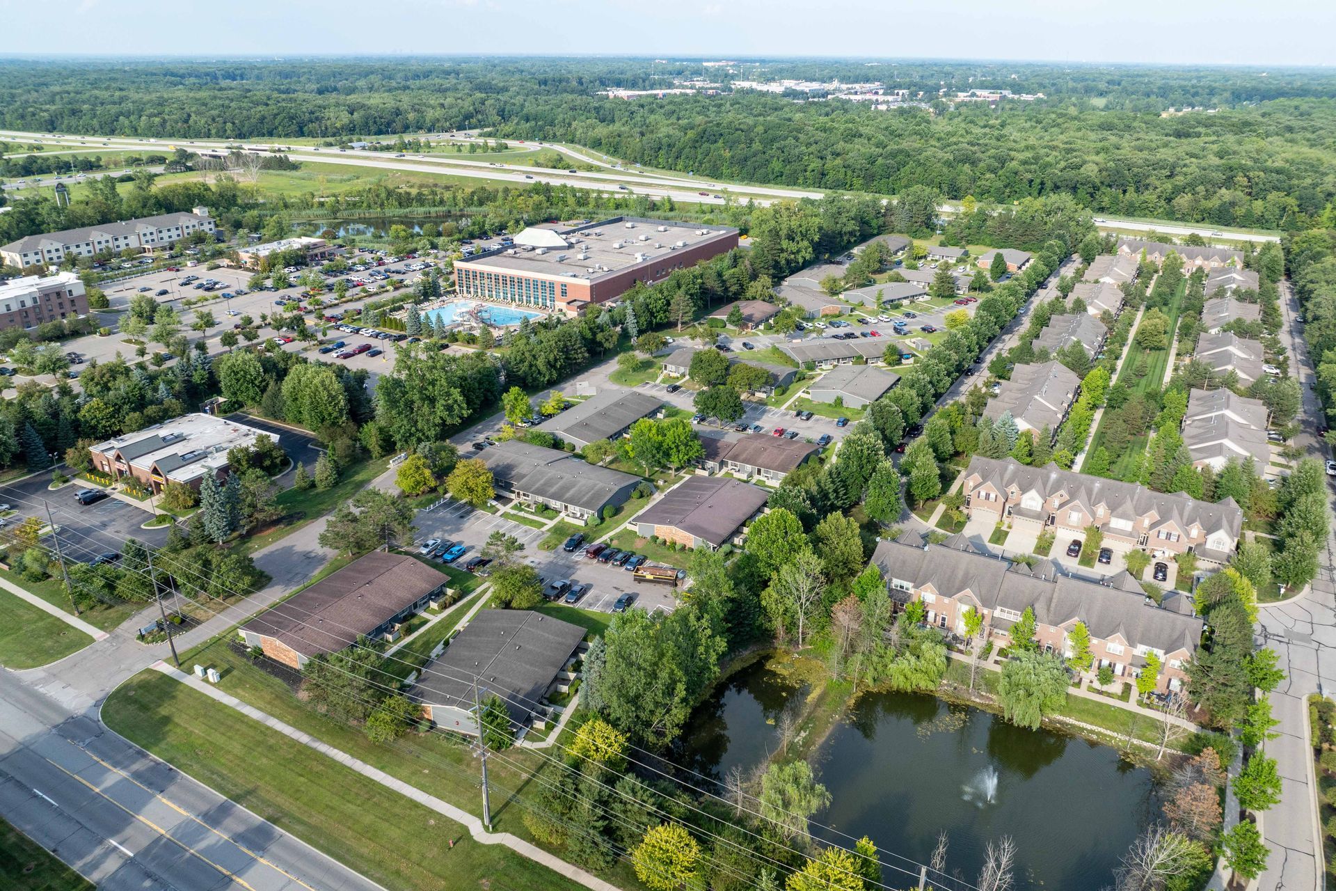 Aerial view of a suburban area with buildings, roads, parking, a pond, and surrounding trees.