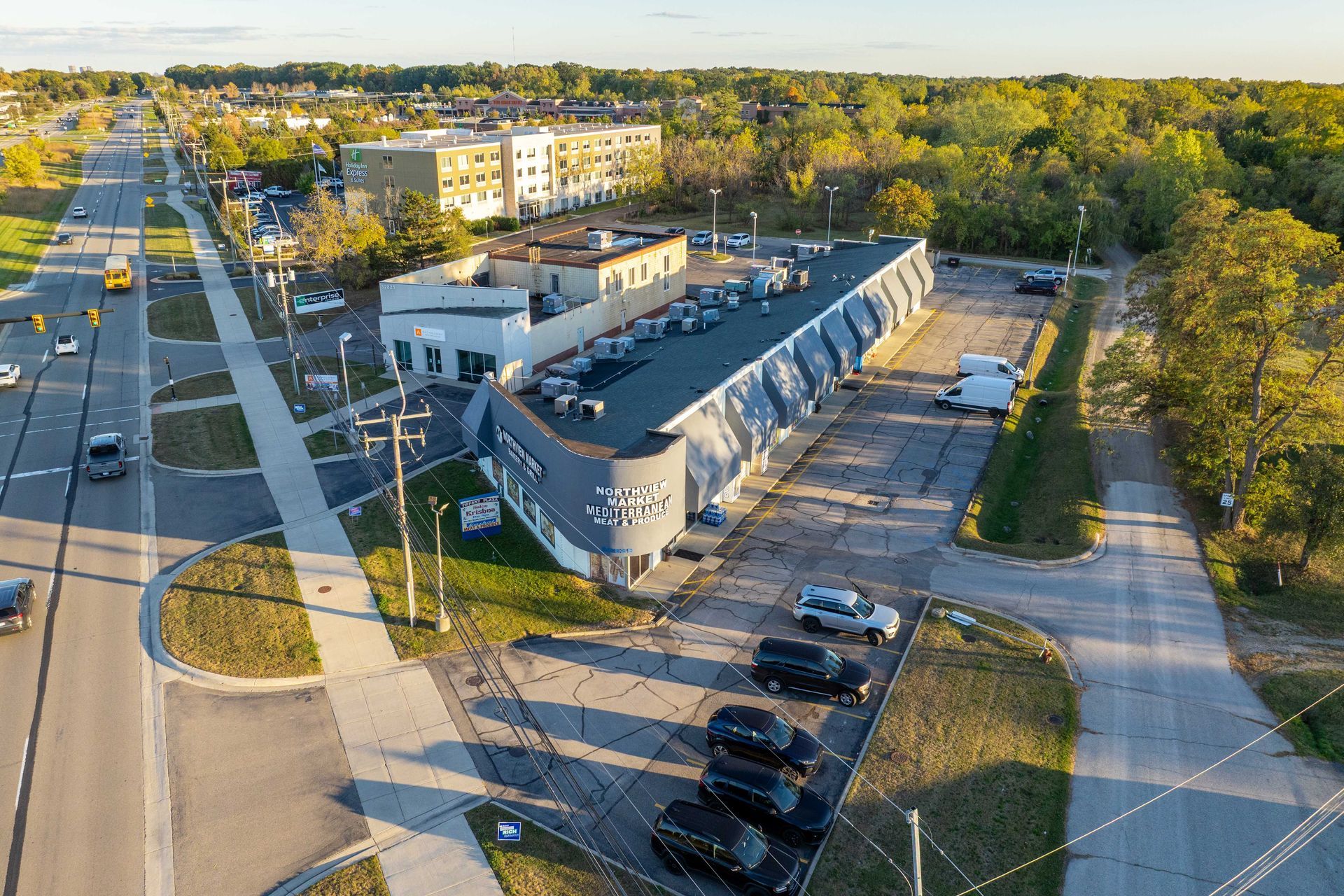 Aerial view of a long, blue commercial building with parked cars, beside a road lined with trees.