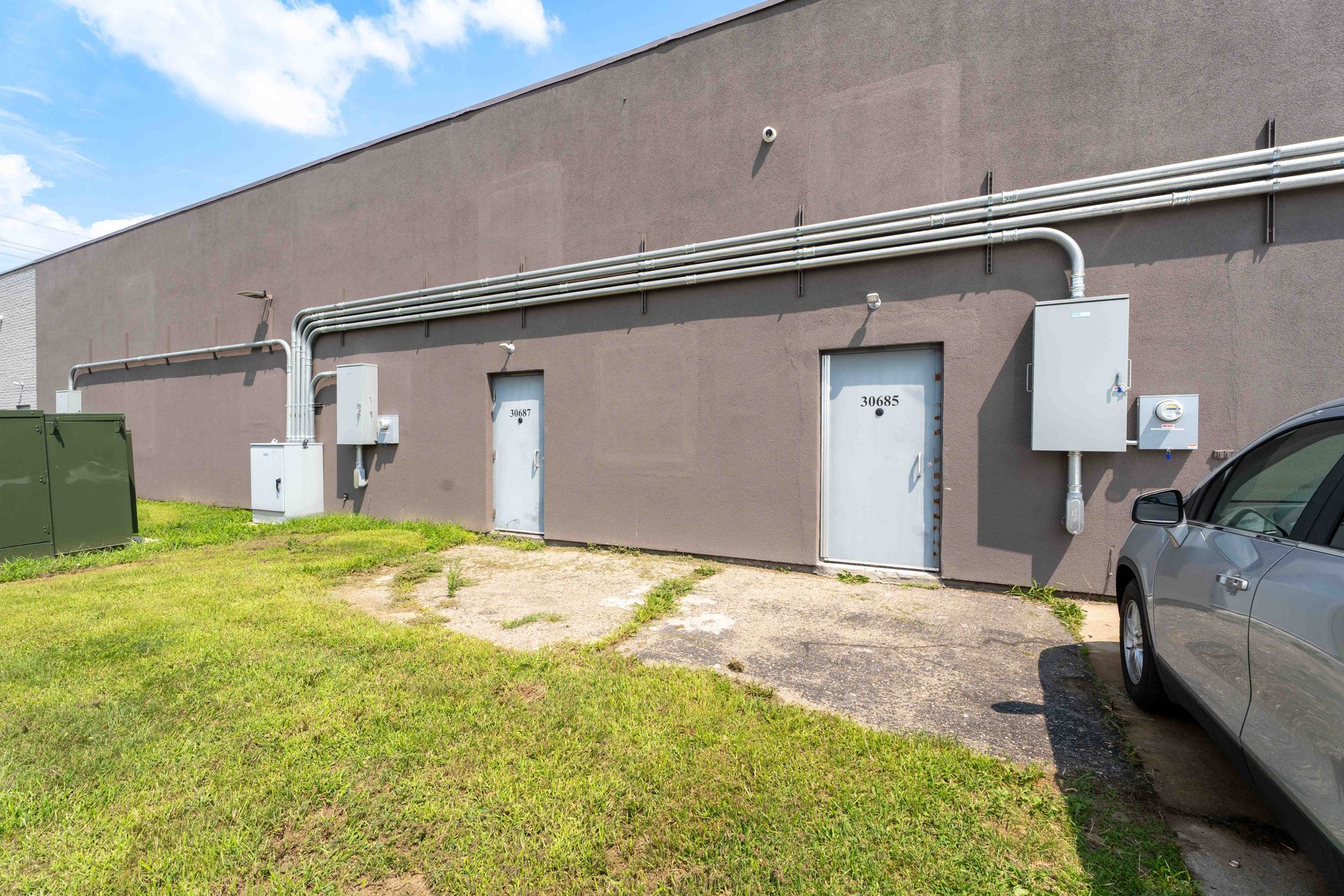 Gray industrial building with doors, electrical boxes, and conduit on a green grassy area.