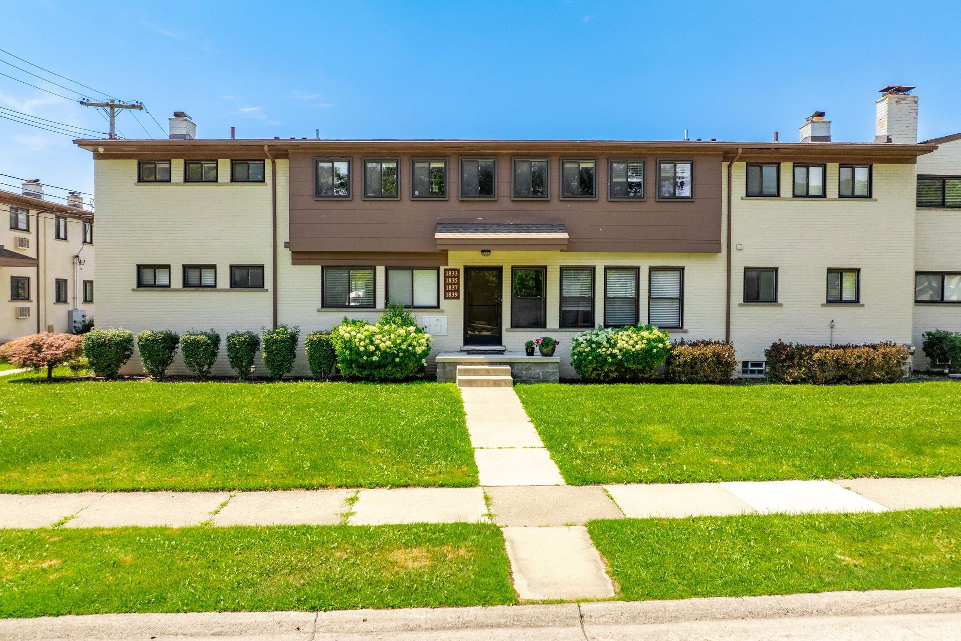 Two-story beige and brown apartment building with green lawn and walkway on a sunny day.