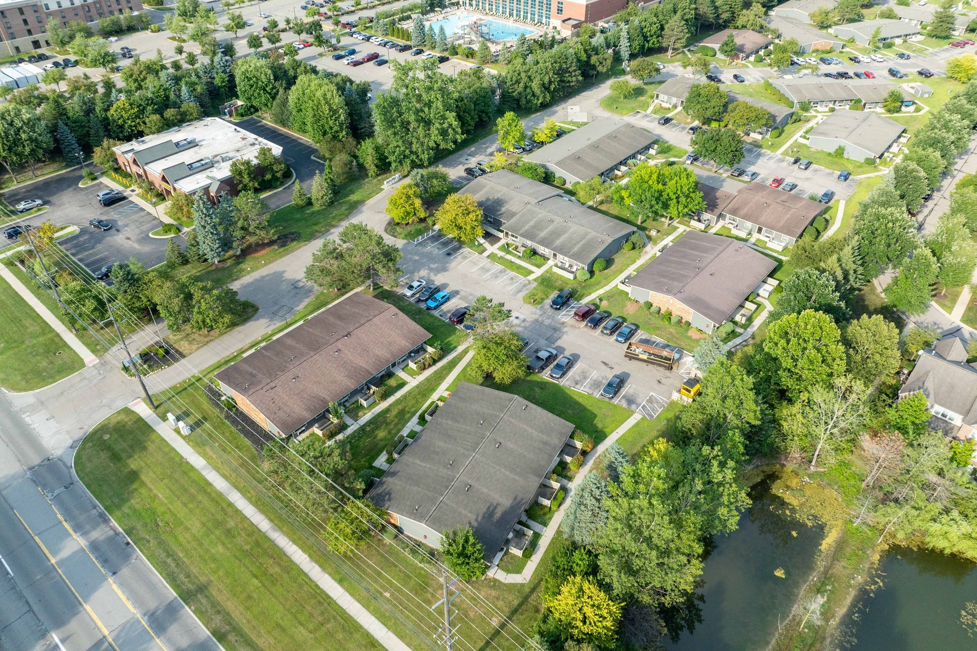 Aerial view of apartment complex with parking, trees, and a pond on a sunny day.