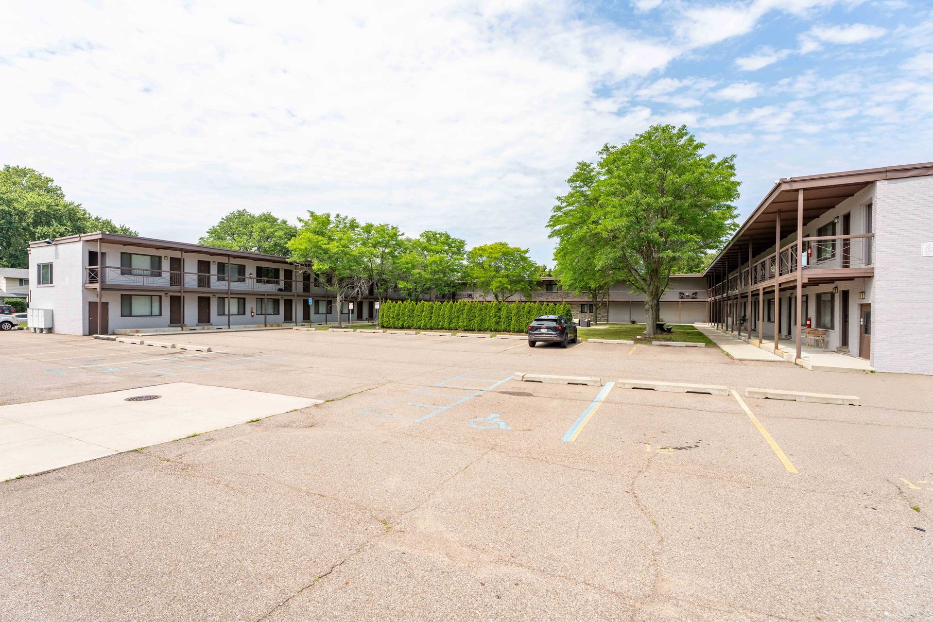 Motel with a large parking lot; buildings are white and brown. Trees are in the background.