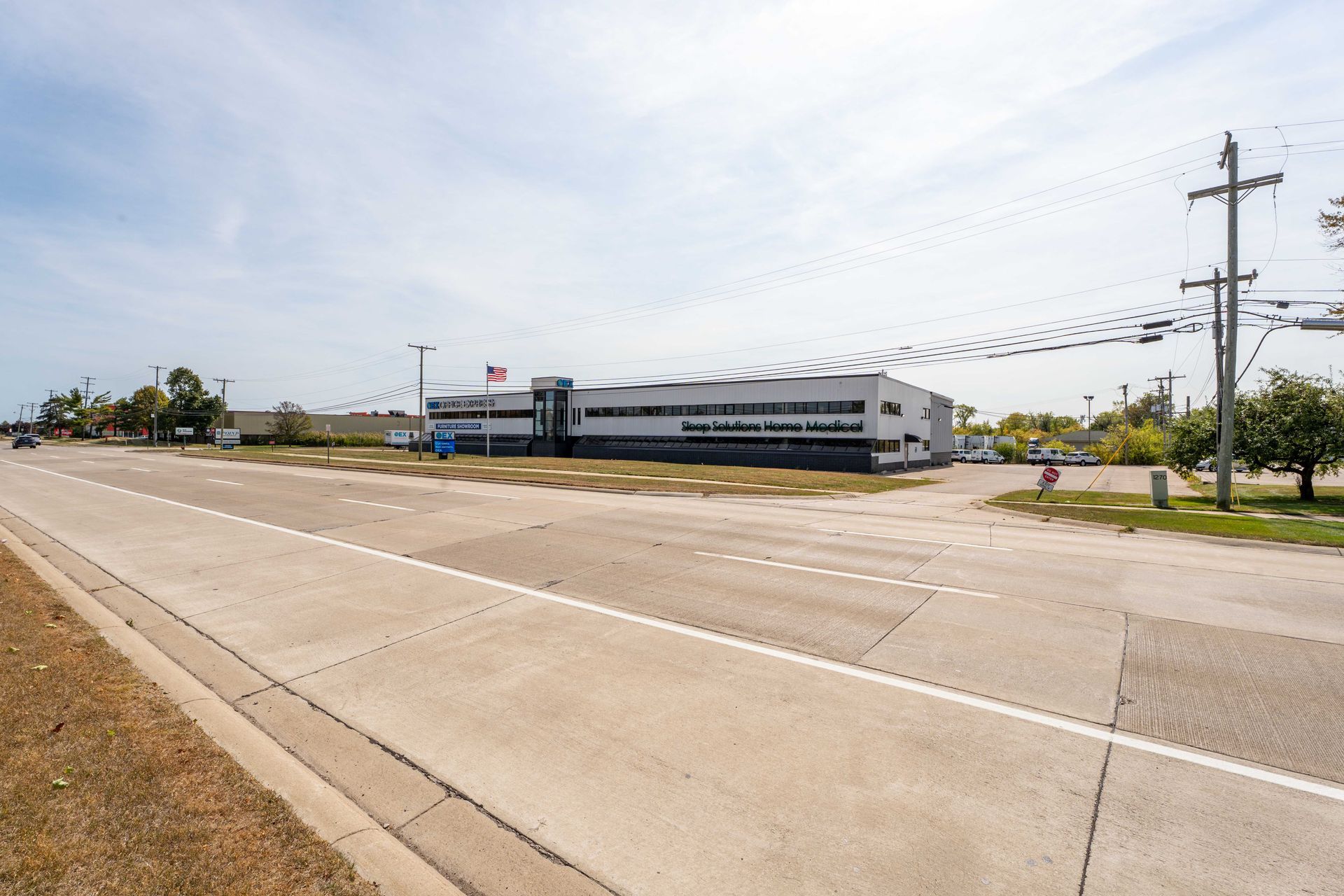 A two-story commercial building with a long front sits along a multi-lane road under a cloudy sky.