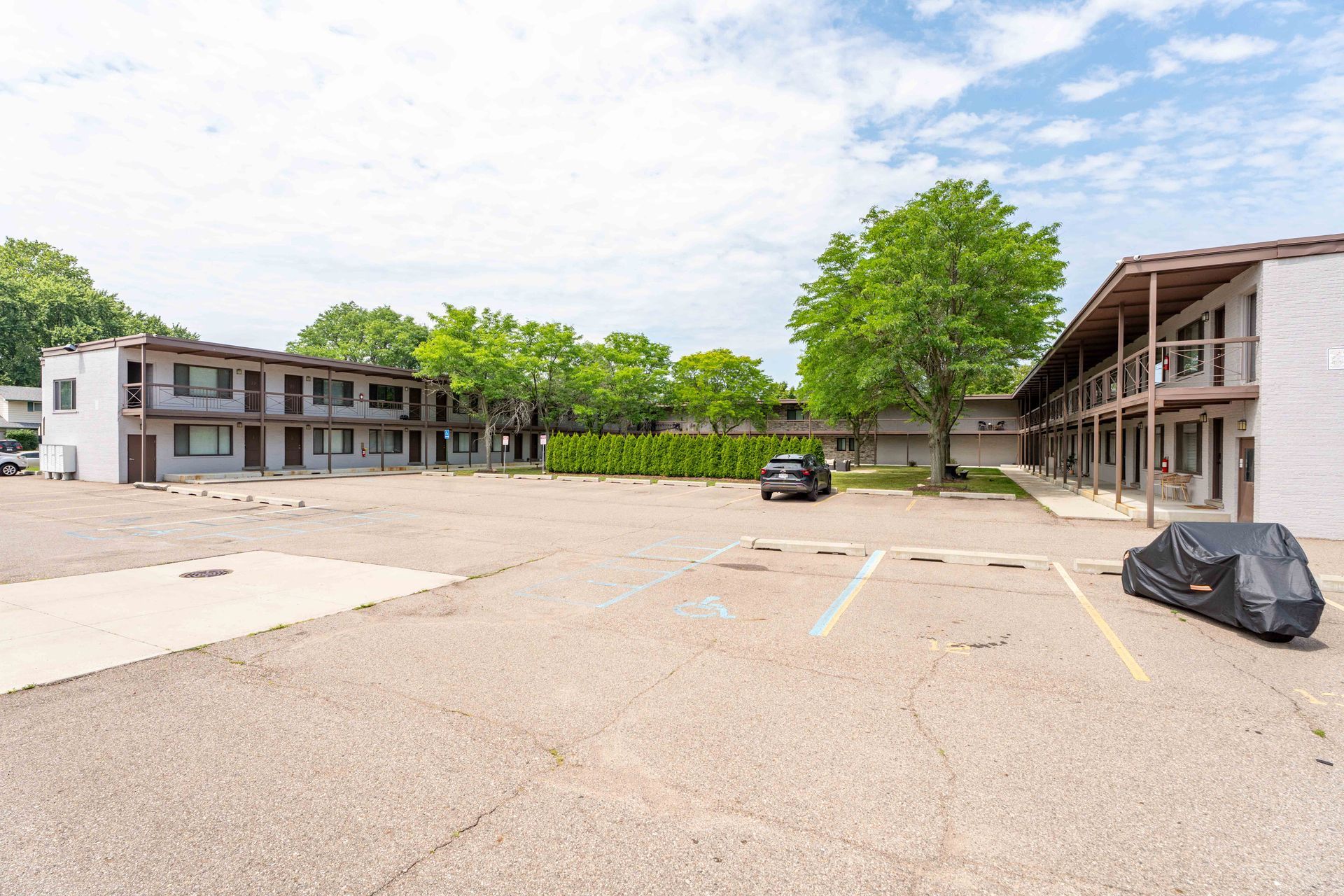 Exterior view of a two-story motel with a large parking lot, blue sky and trees.
