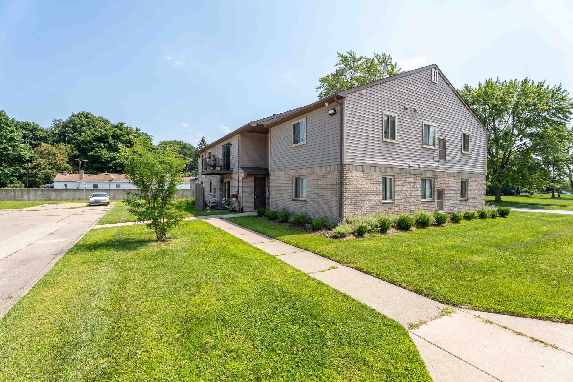 Two-story apartment building with tan siding, brick facade, and green lawn on a sunny day.