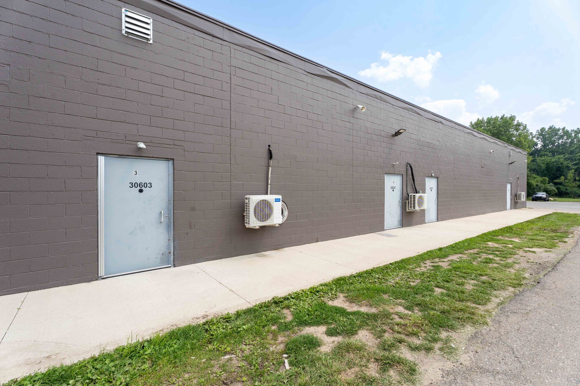 Gray building with three white doors, two with air conditioning units. Sidewalk and green grass in front.
