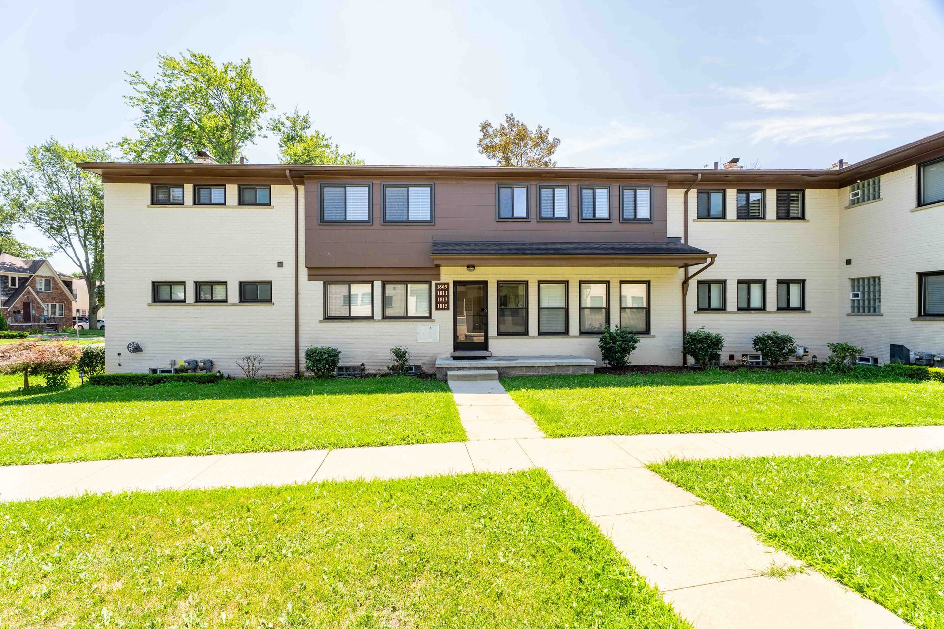 Two-story tan and brown apartment building with green lawn and sidewalk leading to the front door.