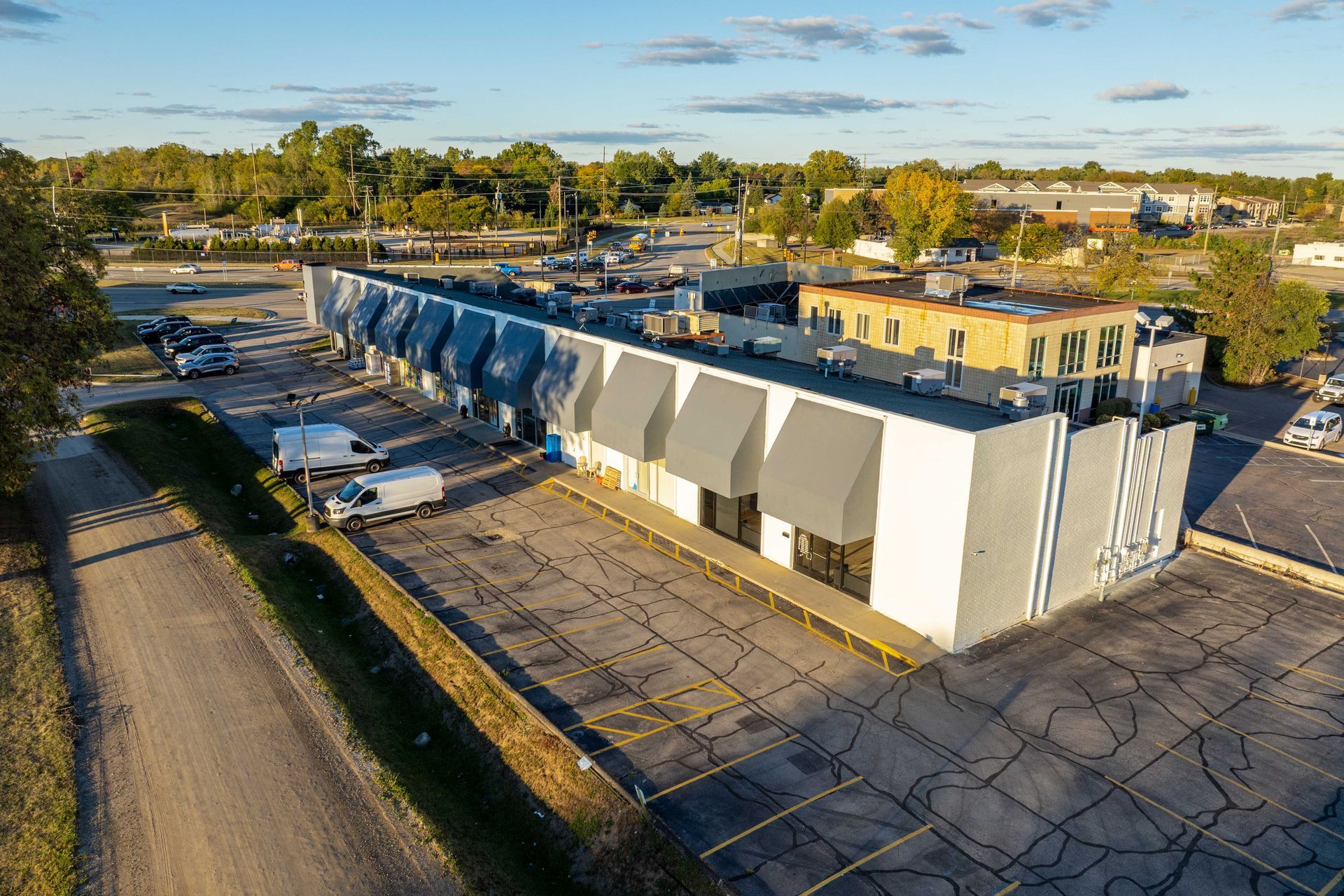 A long, white and gray commercial building with gray awnings, parked cars, and a clear sky.