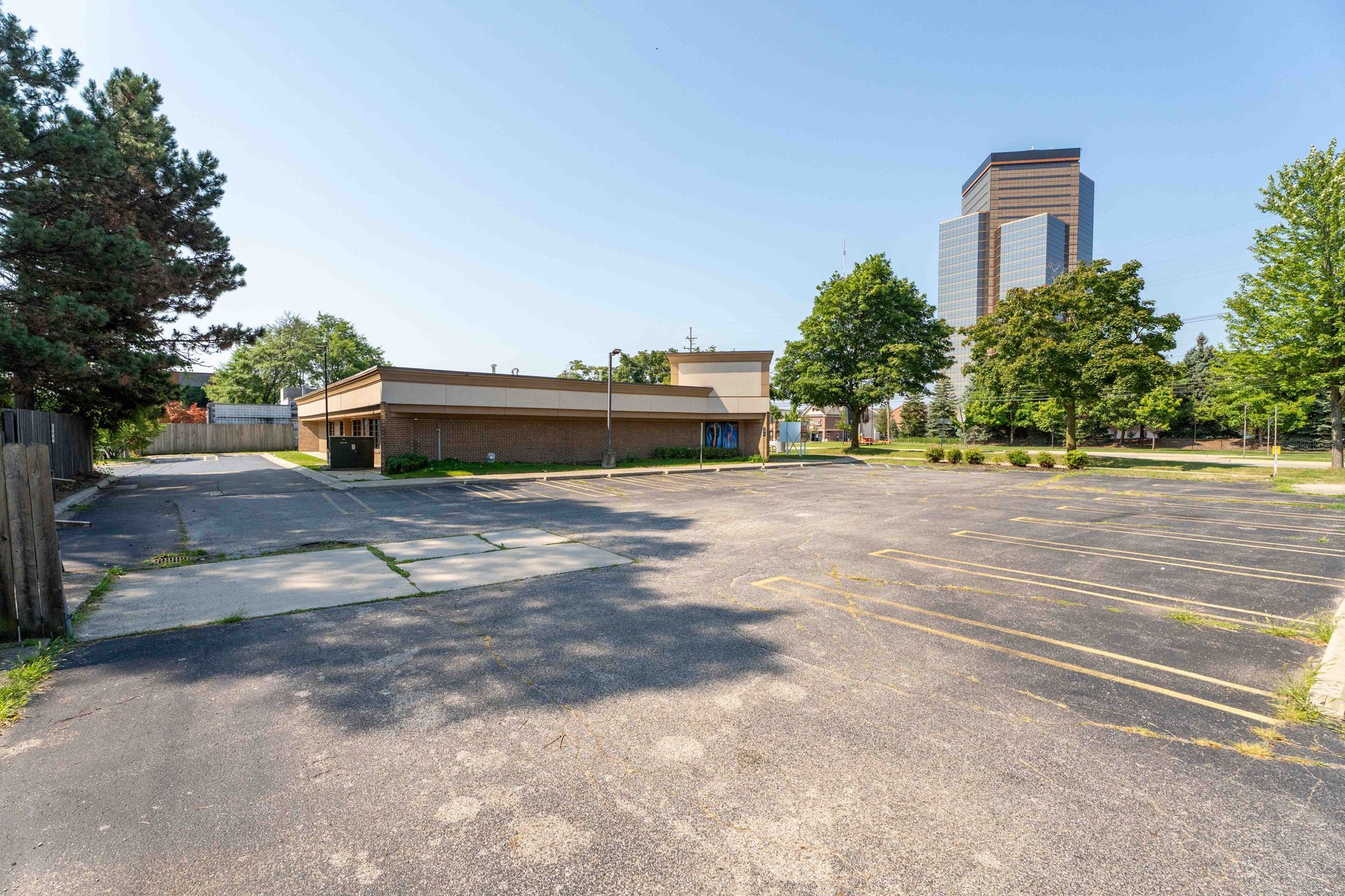 Empty asphalt parking lot with a low, brown building and a tall skyscraper in the background.