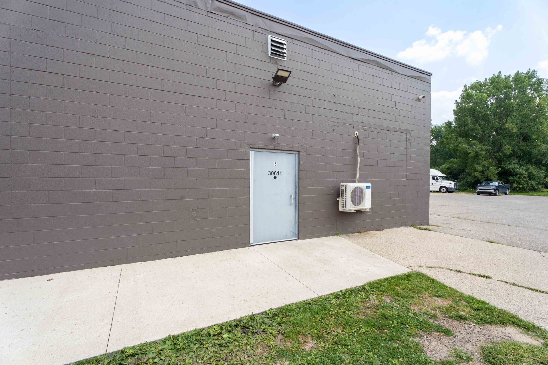 Grey building exterior with white door, sidewalk, and air conditioning unit. Green grass and parking area in view.