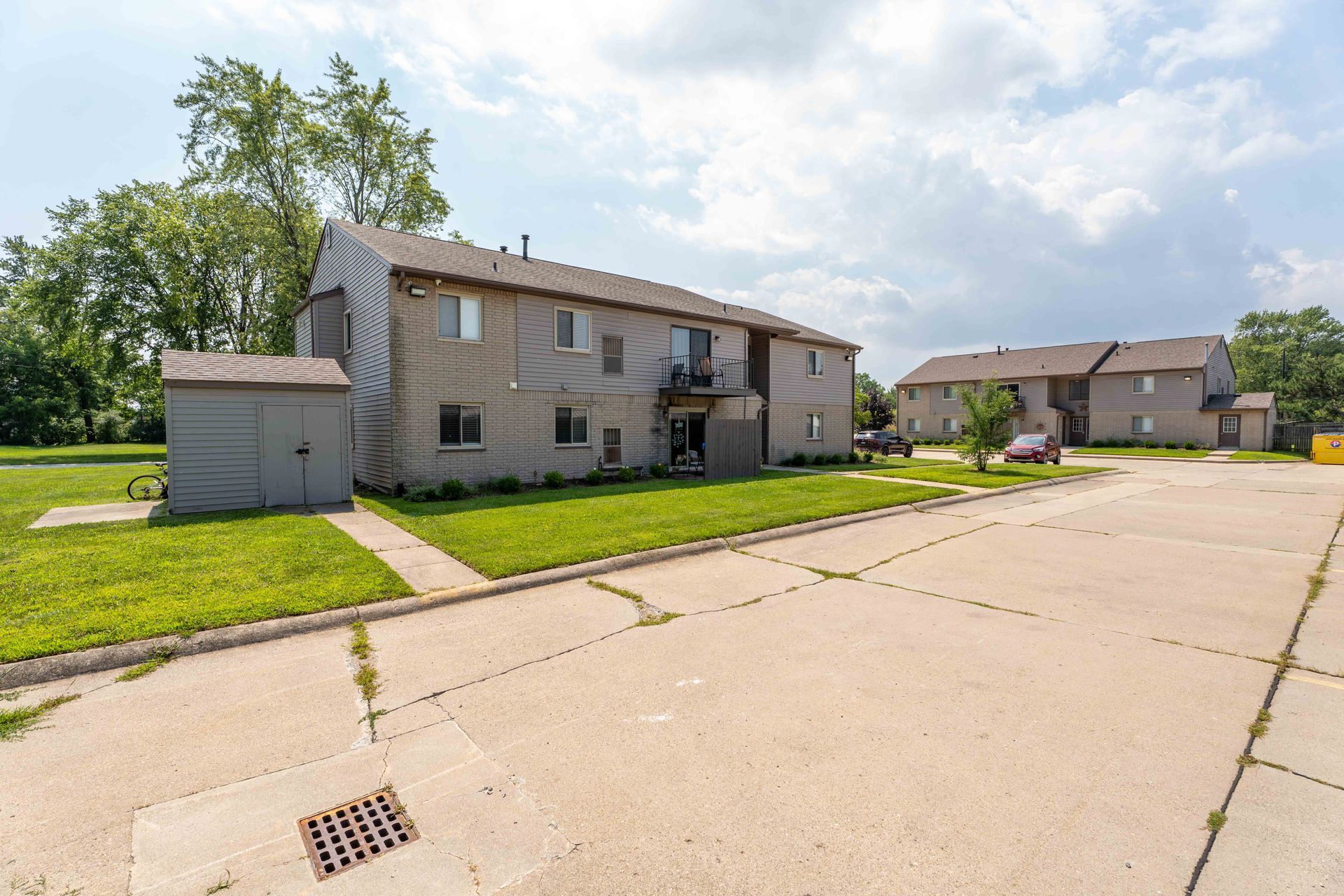 Two-story apartment buildings with grassy lawn and cracked concrete parking area on a cloudy day.