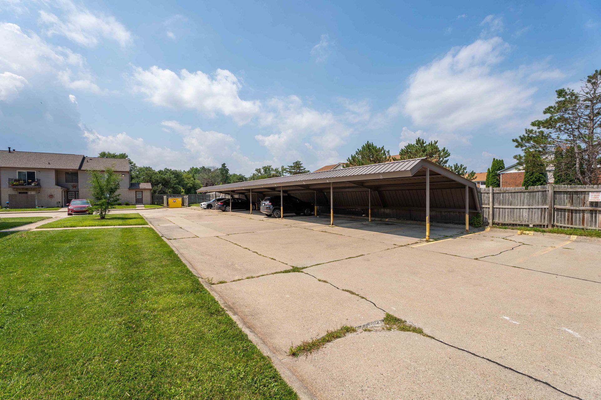 Carport with parked cars on a concrete lot, grassy area, and apartment buildings under a blue sky.