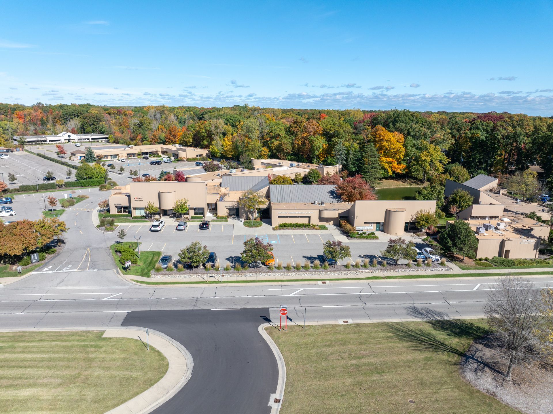 Aerial view of a beige building complex with a parking lot and road in front, surrounded by colorful trees under a blue sky.