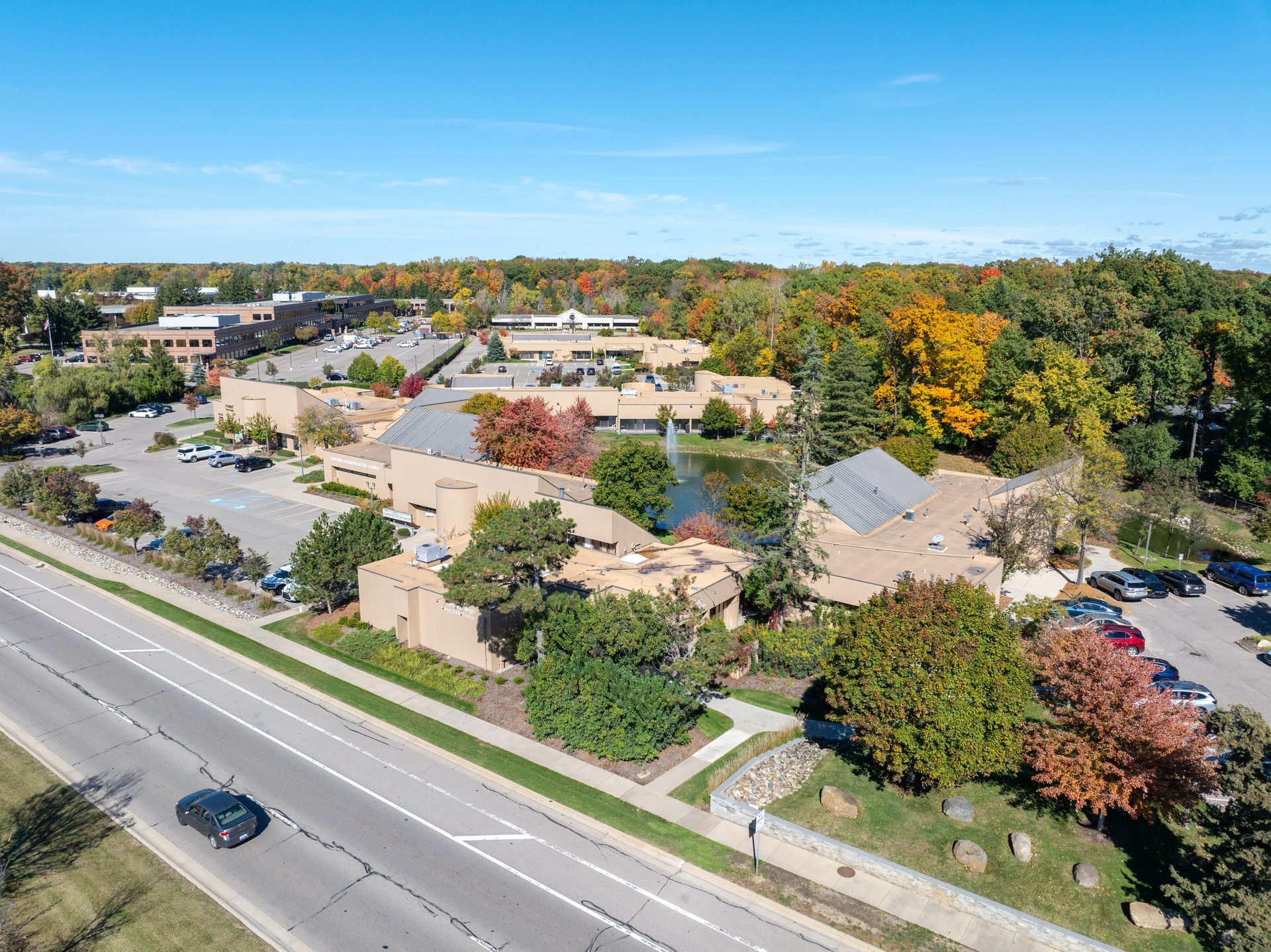 An aerial view of a commercial building with brown roofs, surrounded by trees in fall colors, and a parking lot.