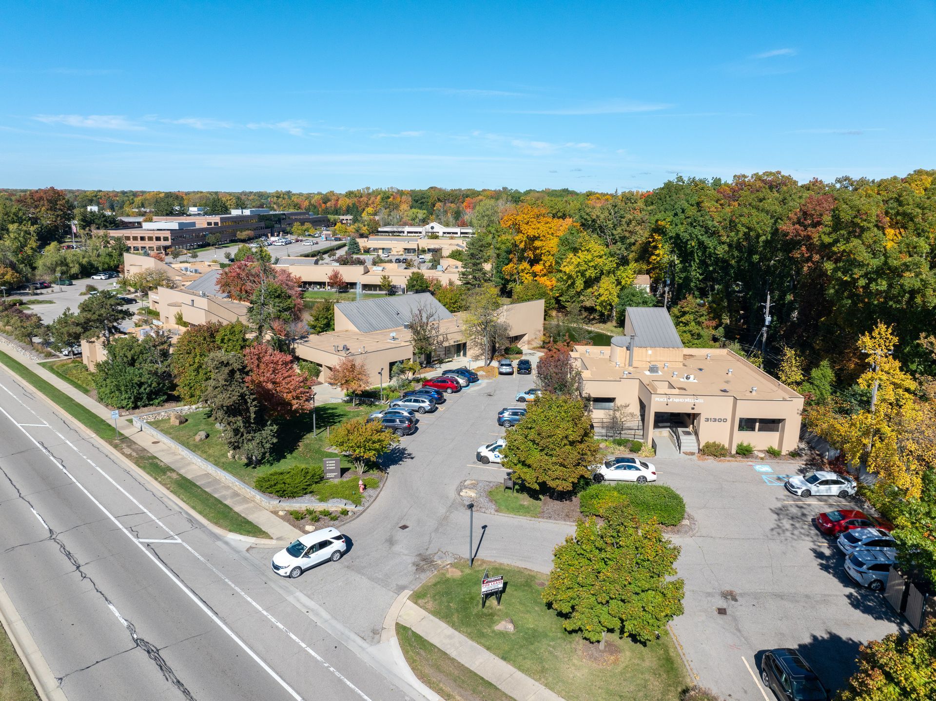 Aerial view of commercial buildings, parking lot with cars, and surrounding trees, clear blue sky.