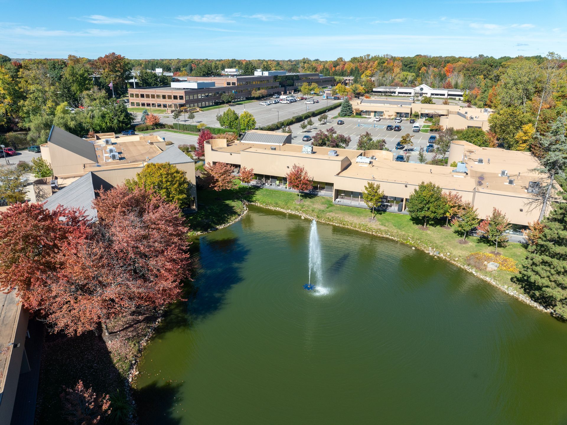 Aerial view of a commercial area with a pond, fountain, and autumn foliage. Buildings and parking lots are visible.